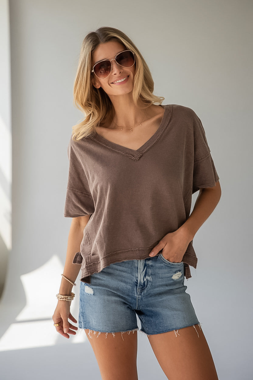 A woman wearing a brown short-sleeve V-neck t-shirt and blue denim shorts, posing in front of a neutral light gray wall.