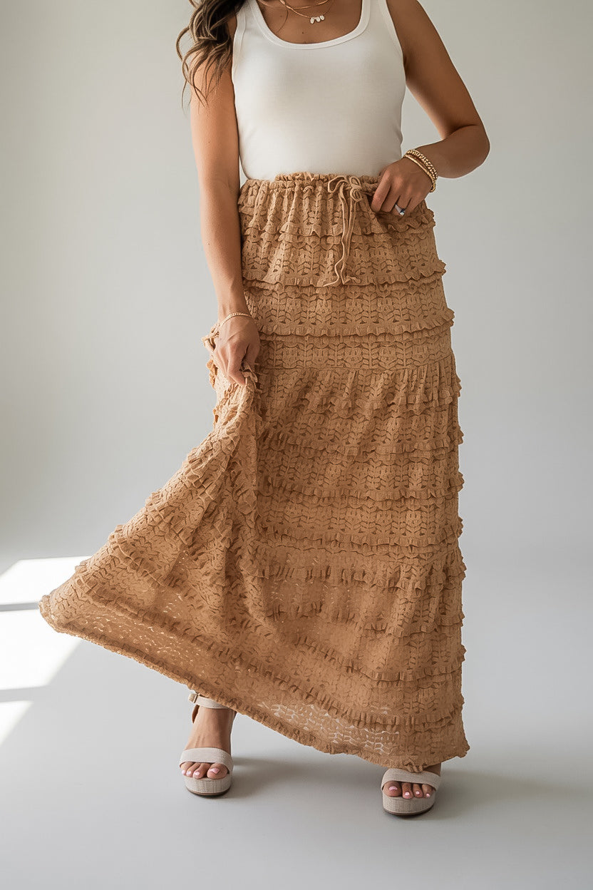A woman wearing a long, tiered, textured tan maxi skirt with a drawstring waist, paired with a white sleeveless top and white heeled sandals.