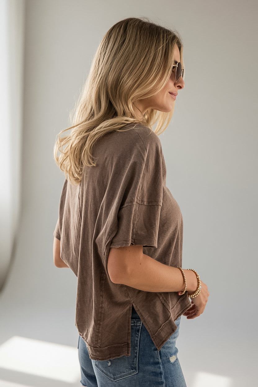 A woman with blonde hair and sunglasses, wearing a brown short-sleeve cropped t-shirt and blue jeans, shown from a side-back profile against a neutral light gray background.