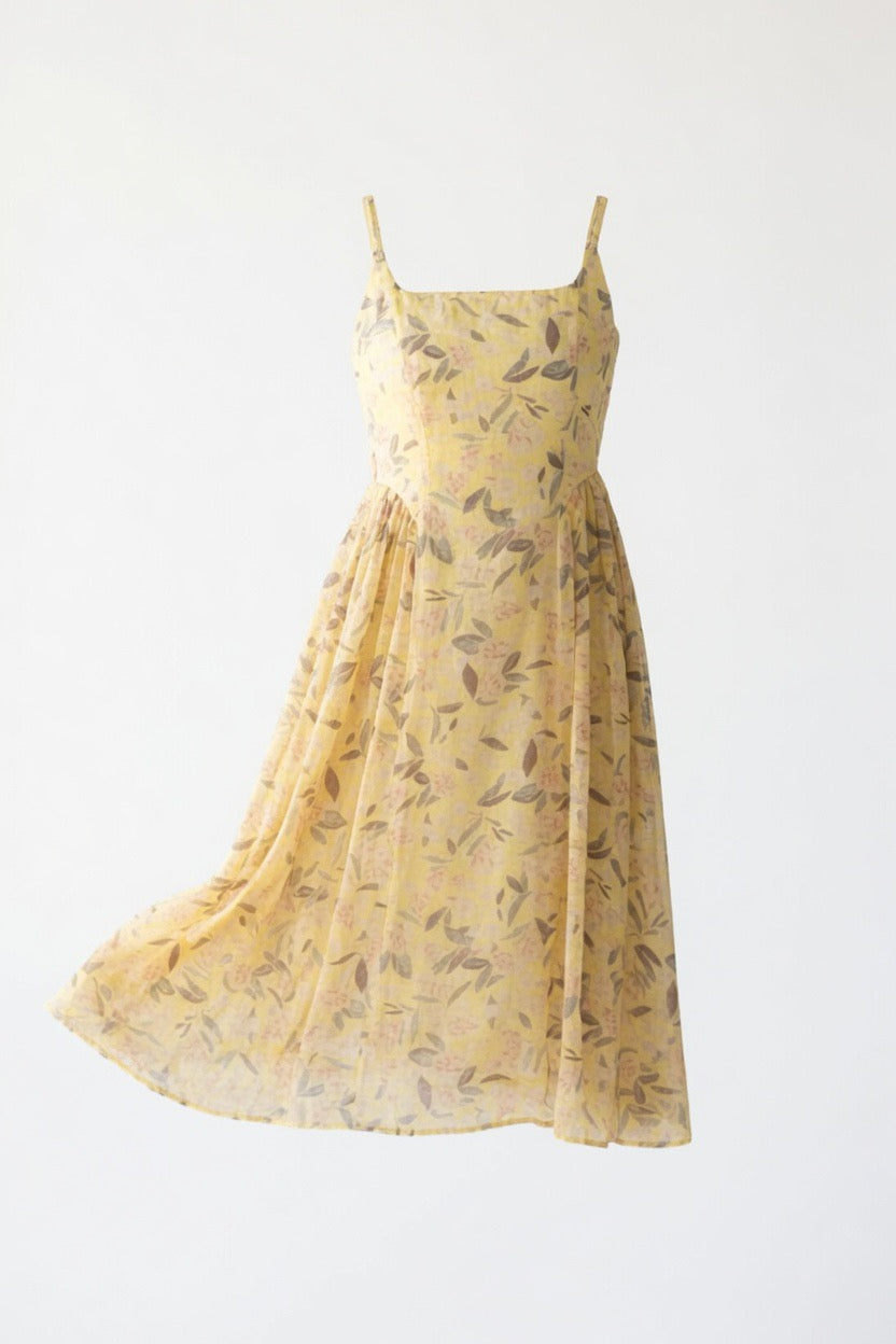 A light yellow sleeveless midi dress with a brown leaf pattern, featuring thin spaghetti straps and a square neckline, shown against a plain white background.
