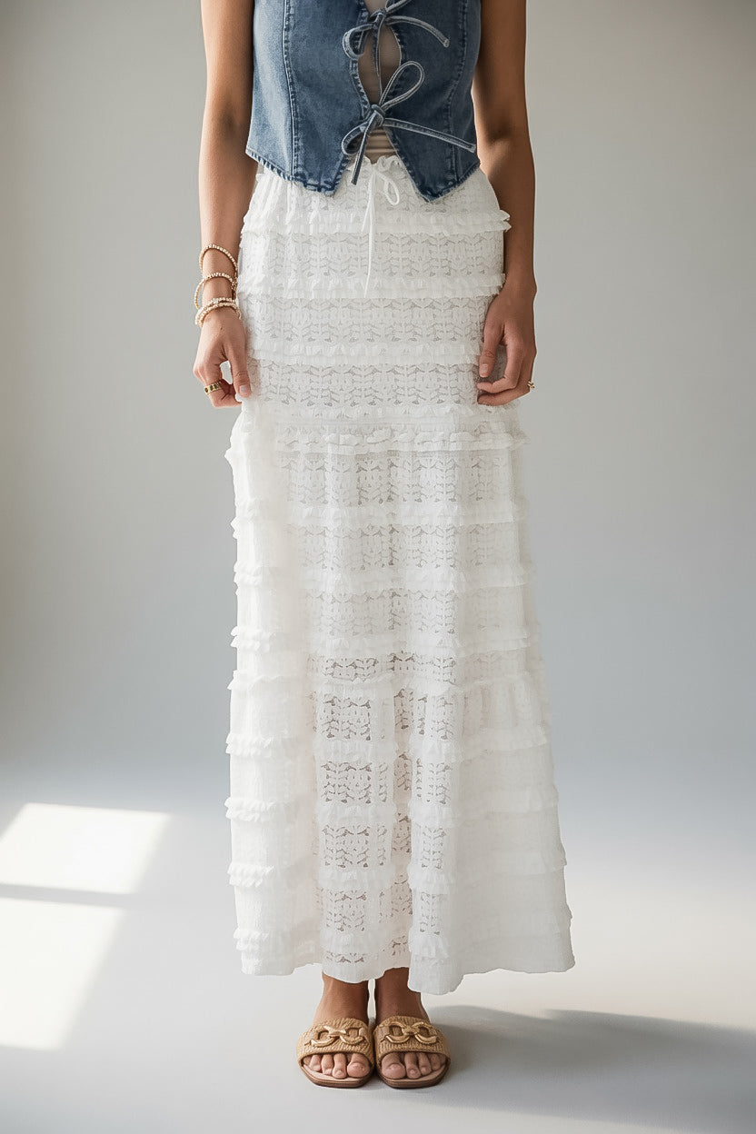 A model wearing a long, tiered white lace maxi skirt paired with a denim corset top and tan slide sandals, standing against a neutral light gray studio background.