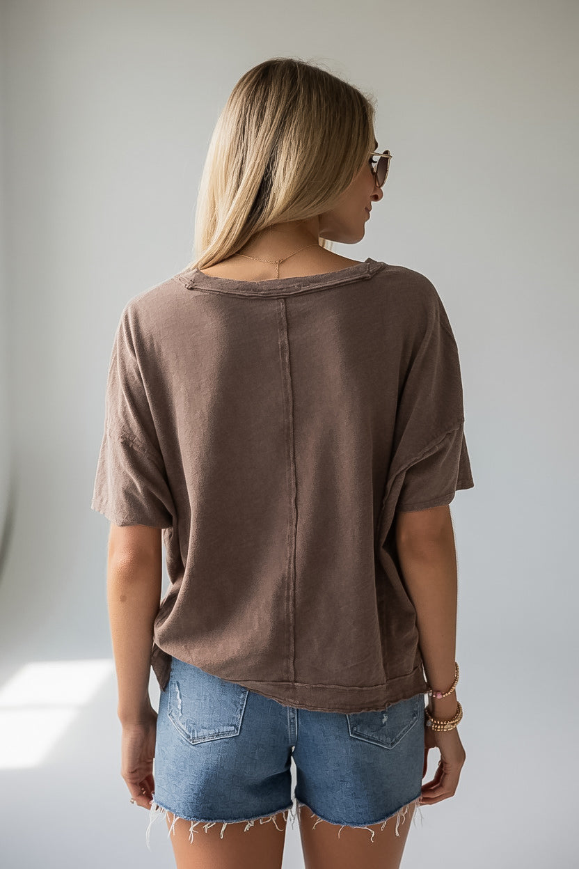 A woman viewed from behind wearing a brown short-sleeve t-shirt with a raw, slightly distressed neckline and denim shorts, standing against a light gray wall.