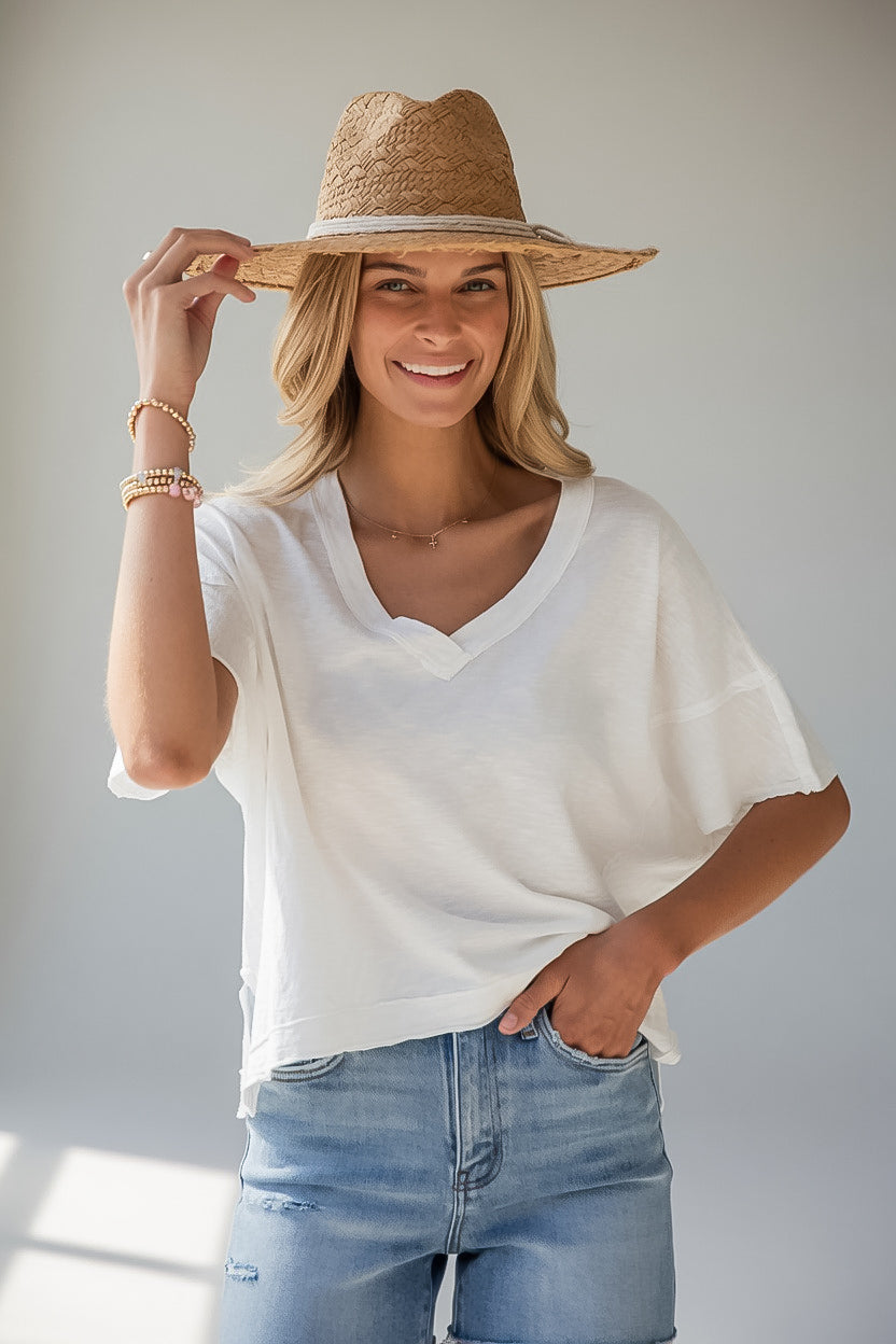 A woman wearing a white short-sleeve V-neck top, light blue jeans, and a straw hat, posing against a light gray background.