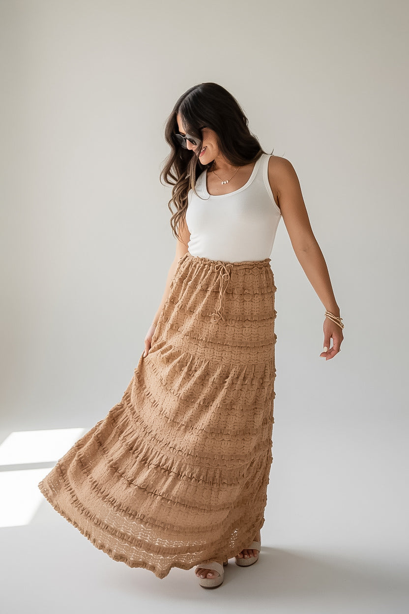 A woman wearing a long, tiered tan maxi skirt with a white ribbed tank top, standing in a studio setting.