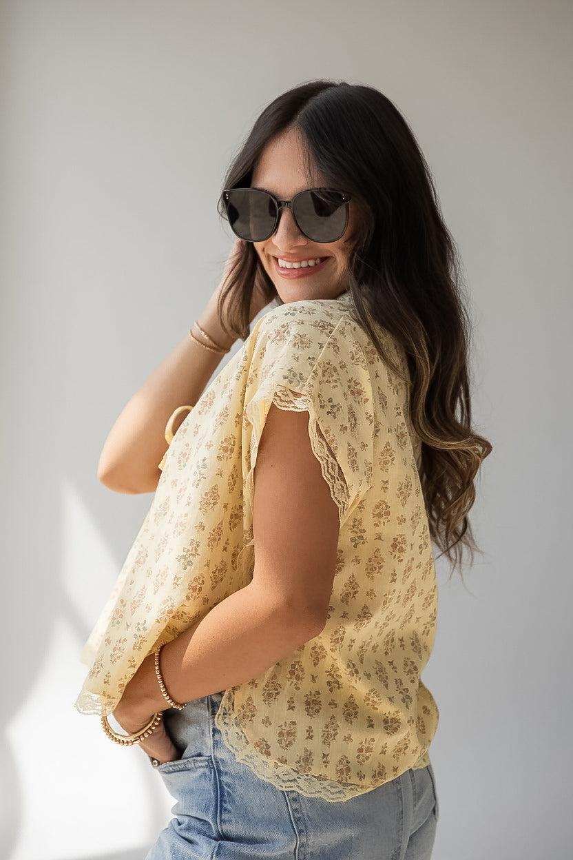 A woman wearing a light yellow sleeveless blouse with a delicate floral pattern and scalloped edges, paired with light blue jeans and dark sunglasses, posing against a neutral light gray background.