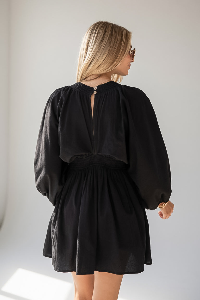 A woman wearing a black long-sleeve mini dress with a gathered waist and puff sleeves, shown from the back in a bright indoor setting.