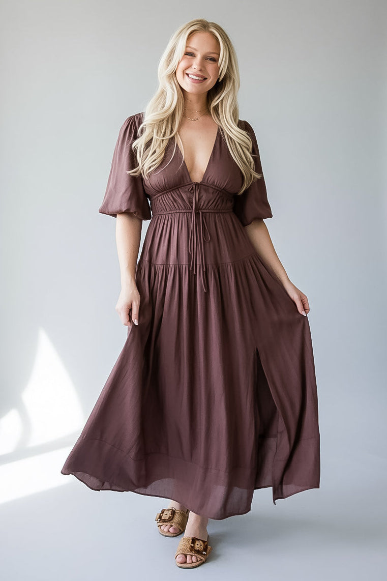 A woman modeling a long, flowy midi dress in a brown color, standing against a light gray studio background.