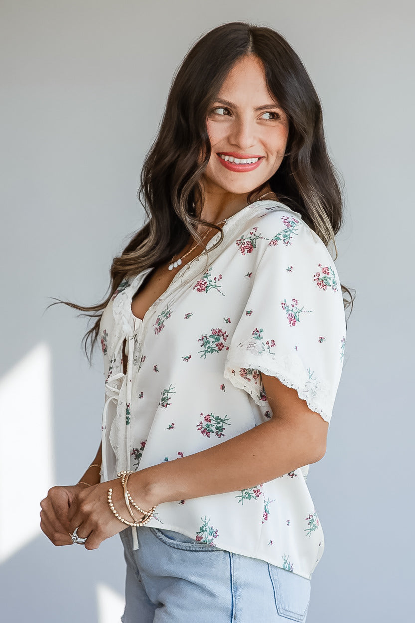 A woman wearing a short-sleeve white blouse with a delicate pink and green floral print, paired with light blue jeans, posing against a neutral light gray background.