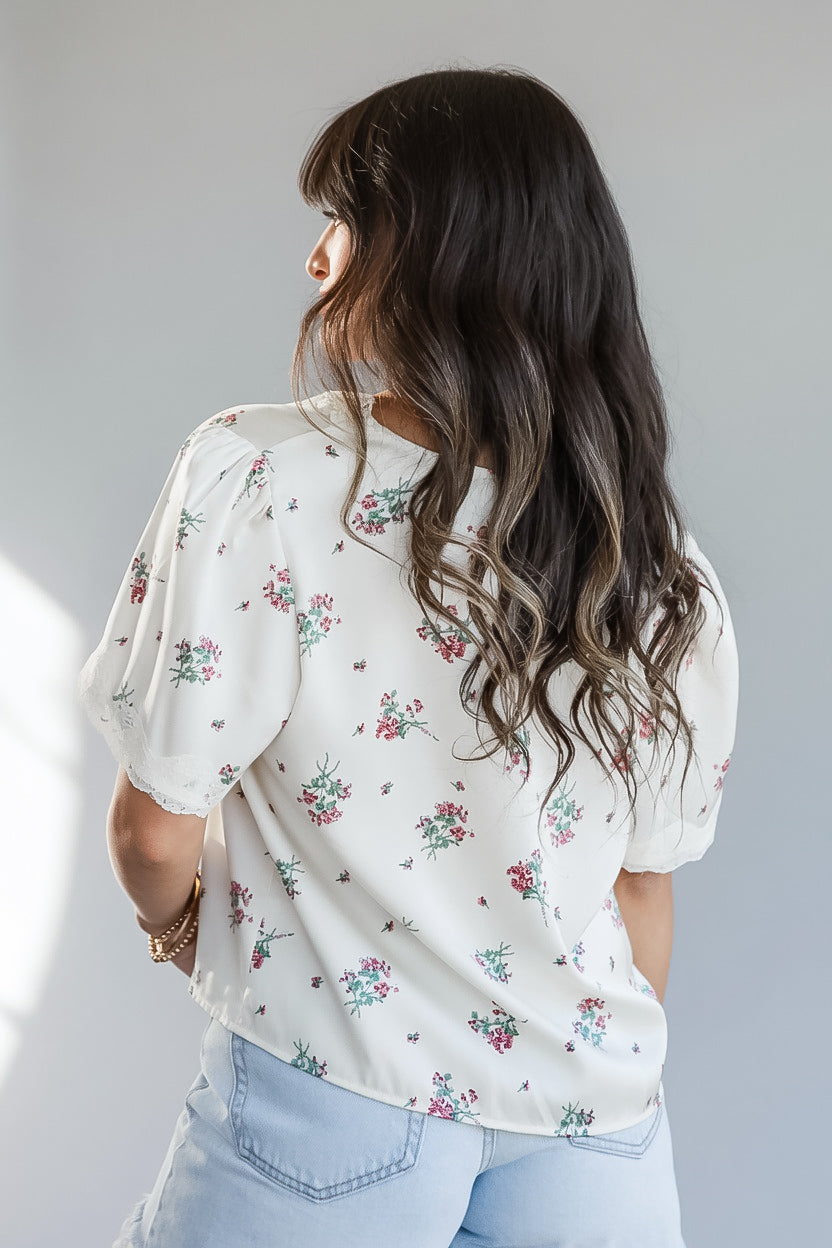 A woman viewed from behind wearing a cream-colored short-sleeve blouse with a delicate pink and green floral print, paired with light blue jeans, against a plain light gray background.