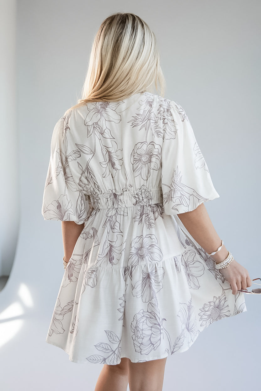A woman viewed from behind wearing a short white tiered dress with a gray floral line print, featuring flutter sleeves and a gathered waist.