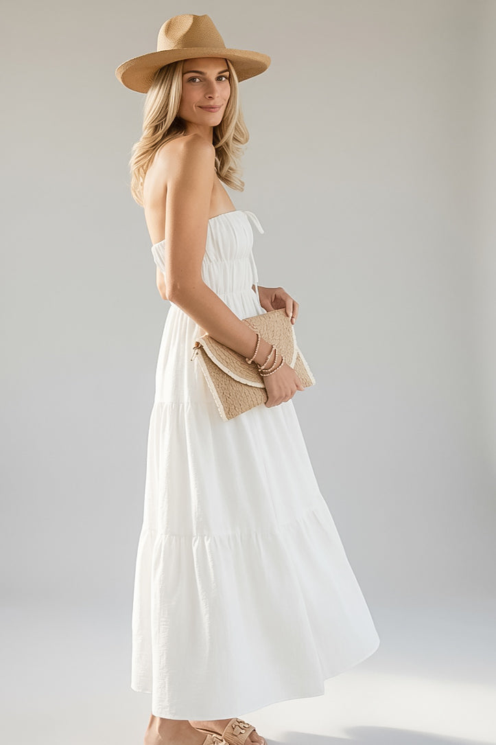 A woman modeling a strapless white tiered maxi dress, styled with a tan wide-brim hat, a woven tan clutch, and tan sandals against a light gray studio background.