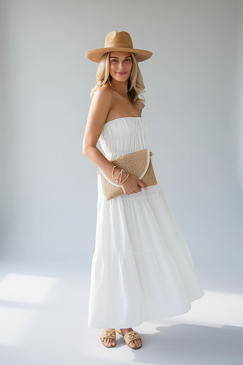 A woman modeling a white strapless maxi dress with a tiered skirt, paired with a tan wide-brim straw hat, a woven clutch, and tan slide sandals, set against a light gray studio background.