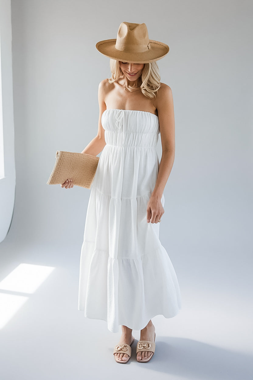 A woman modeling a white strapless tiered maxi dress with a smocked bodice, styled with a wide-brimmed tan hat, a woven clutch, and tan sandals in a bright white studio setting.
