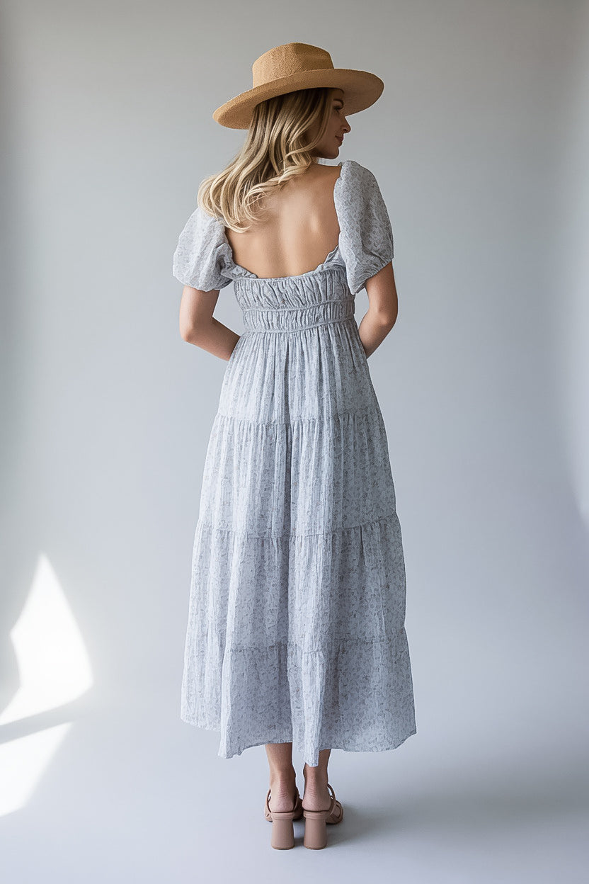 A woman viewed from behind wearing a light gray and white patterned tiered maxi dress with puff sleeves and a smocked back, paired with a tan wide-brim hat and beige heels, set against a plain light gray studio background.