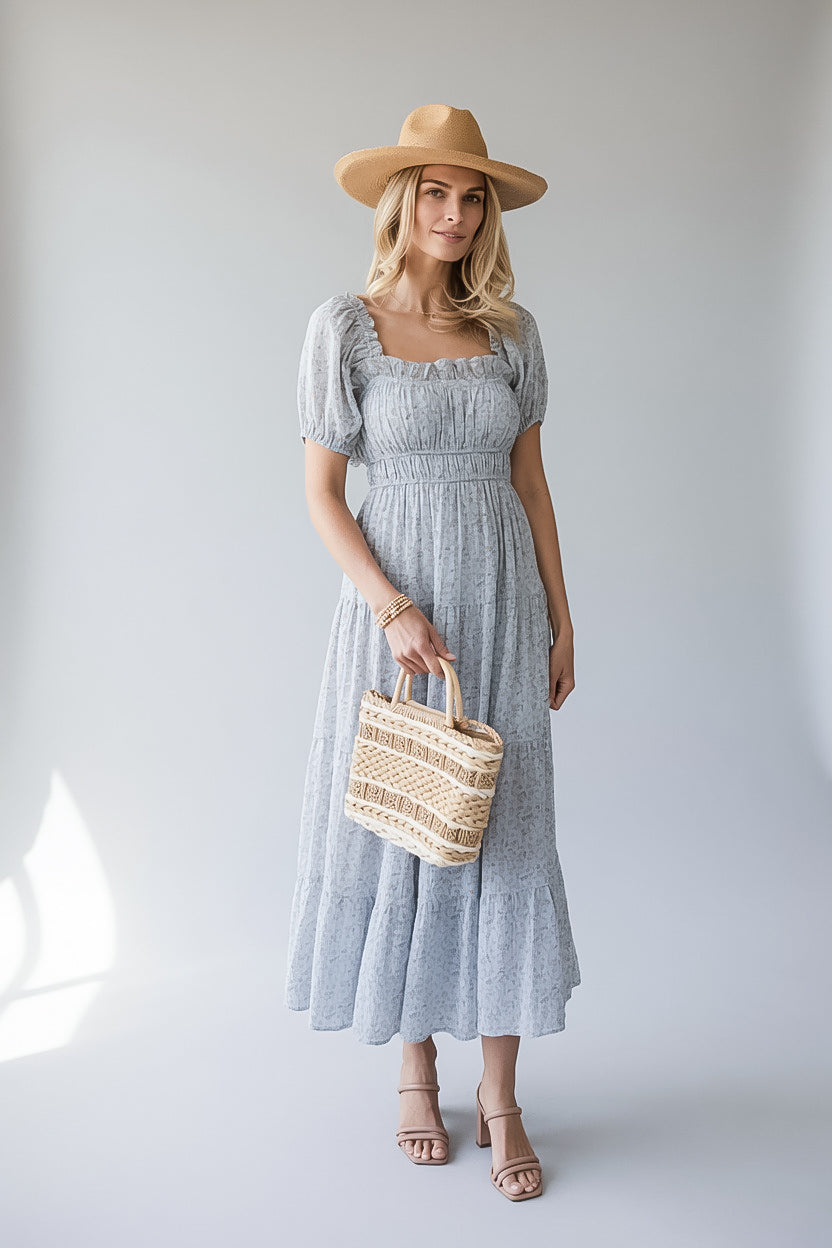A woman modeling a light blue-gray smocked midi dress with puff sleeves, holding a woven straw tote bag and wearing a tan wide-brim hat and heeled sandals against a light gray studio background.