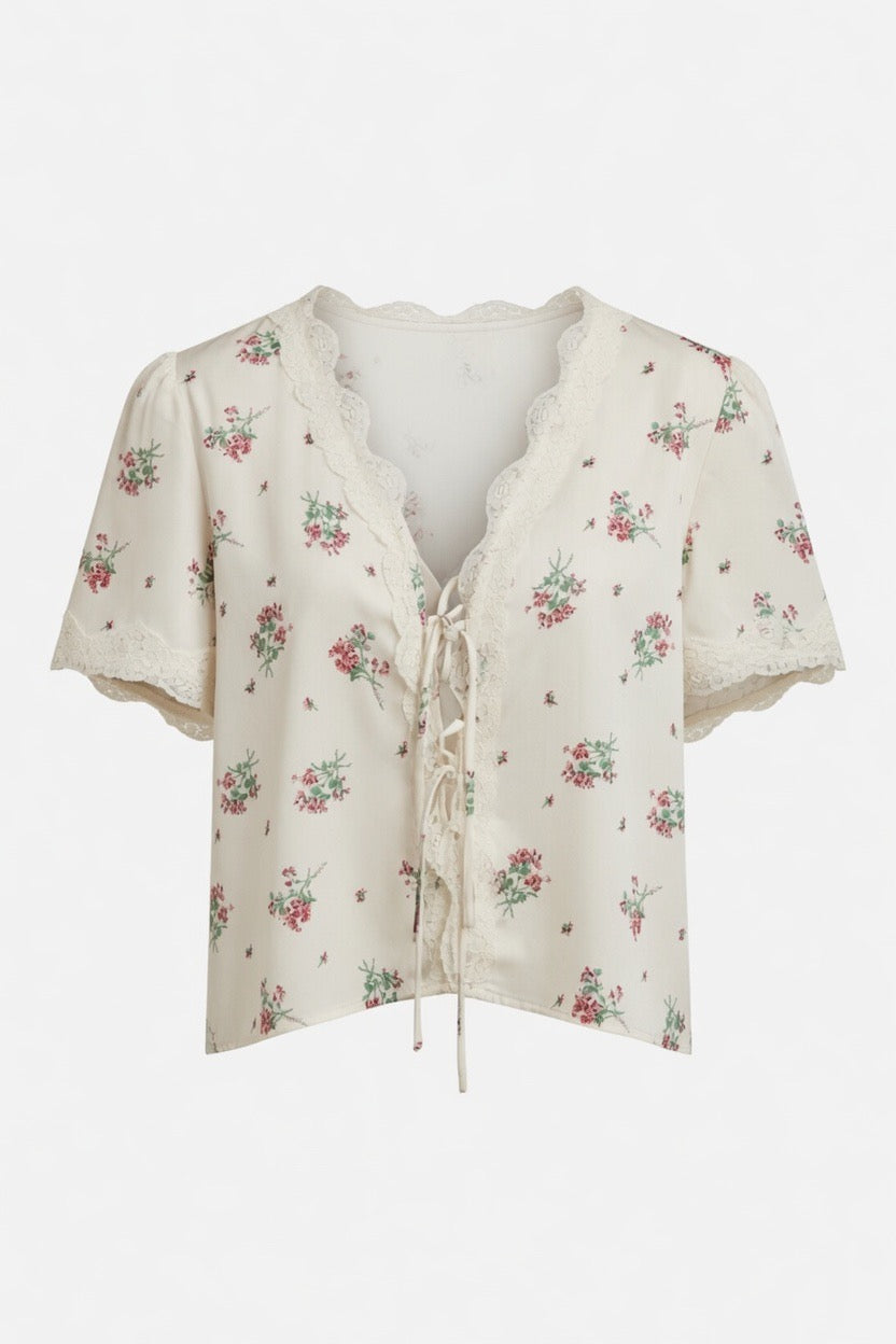 An off-white short-sleeve blouse with a delicate pink and green floral print, featuring a lace-trimmed V-neckline and front tie closure, shown against a plain white background.