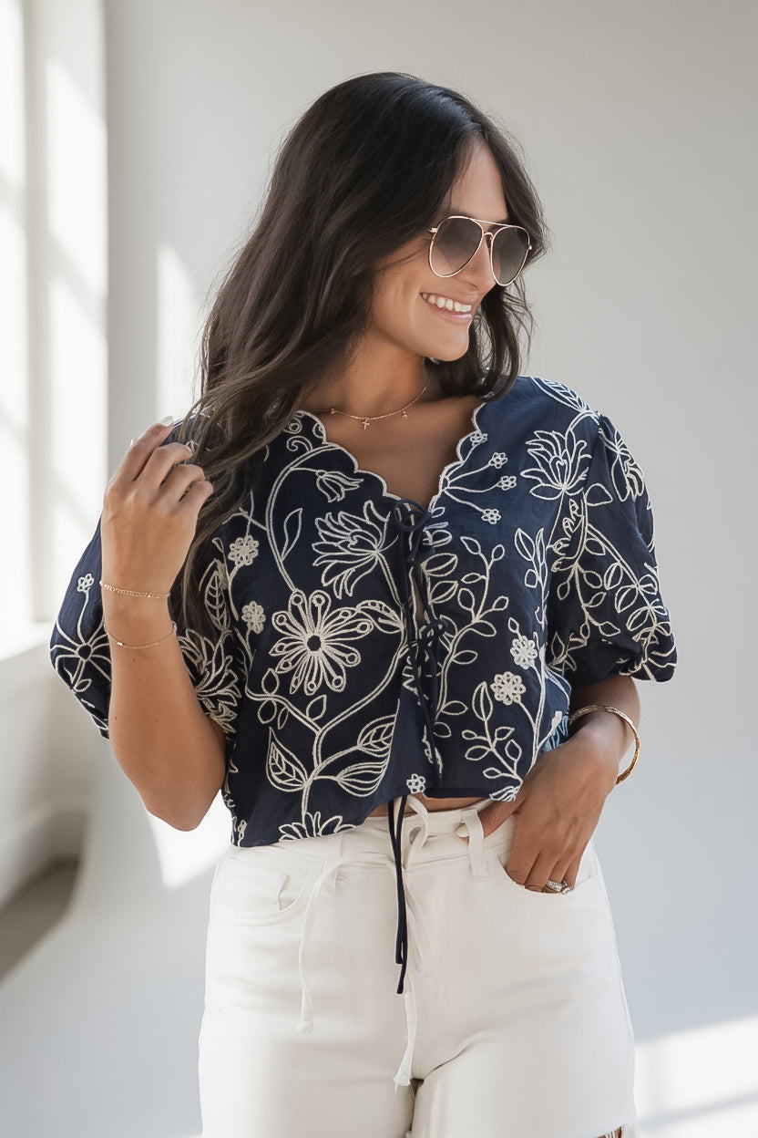 A woman wearing a navy blue short-sleeve blouse with a white floral line pattern, paired with white drawstring shorts and sunglasses, standing in a brightly lit indoor setting.