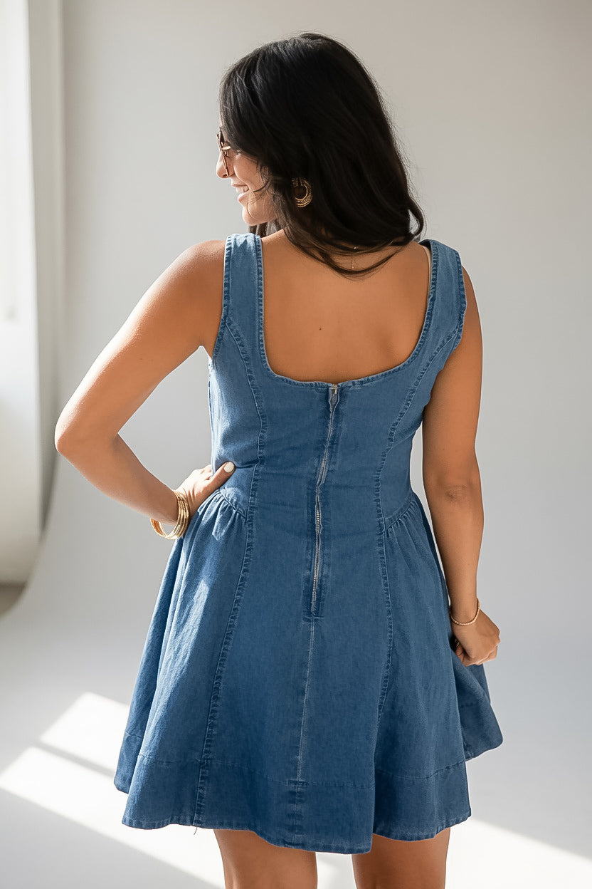 A woman wearing a blue denim sleeveless dress, photographed from behind to show the back zipper and fit, standing in a bright indoor setting.