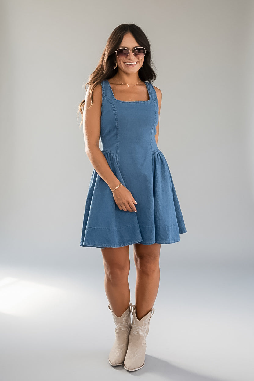 A woman modeling a sleeveless blue denim-like mini dress with a square neckline and flared skirt, paired with light beige cowboy boots, against a plain light gray studio background.