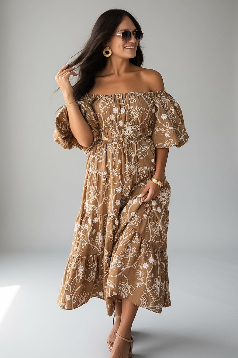 A woman modeling an off-the-shoulder midi dress with puff sleeves and an allover white floral print on a warm tan background, photographed against a light gray studio backdrop.