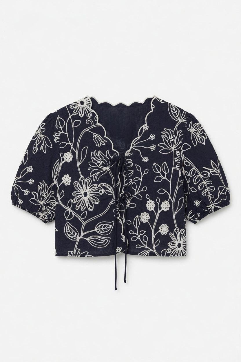 A cropped short-sleeve blouse in navy blue with a white floral line-art pattern, featuring a ruffled V-neckline and a drawstring tie at the front, laid flat against a light gray background.