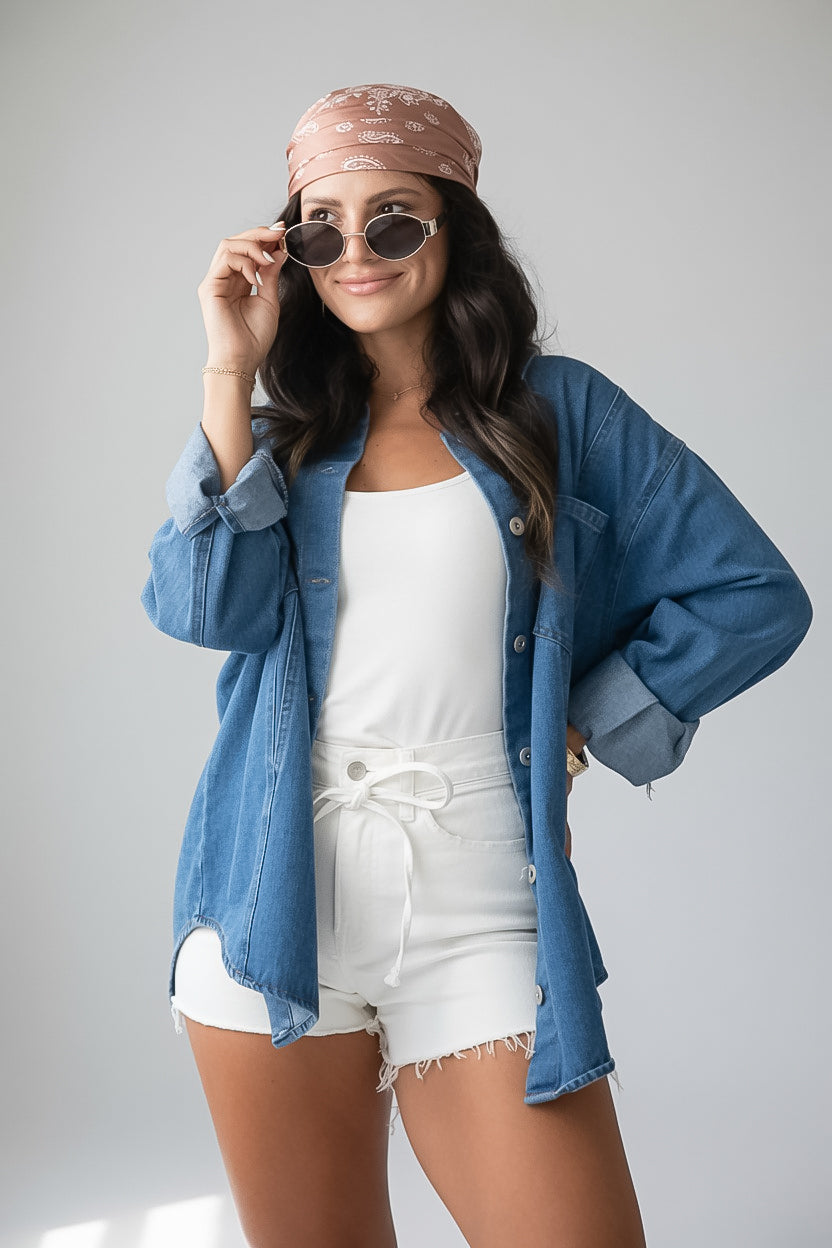 A woman posing in a styled outfit featuring a blue denim button-up shirt, white tank top, white shorts, a pink patterned headband, and sunglasses against a light gray studio background.