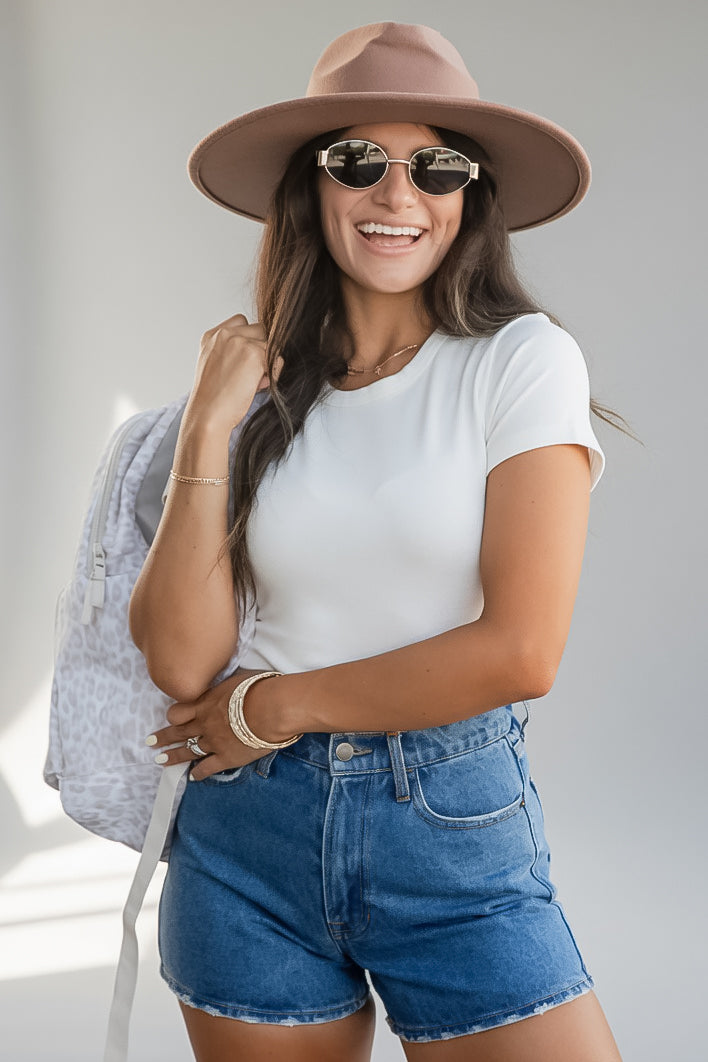 A woman wearing a white short-sleeve t-shirt, blue denim shorts, a brown wide-brim hat, and sunglasses, carrying a light gray patterned backpack against a light gray background.