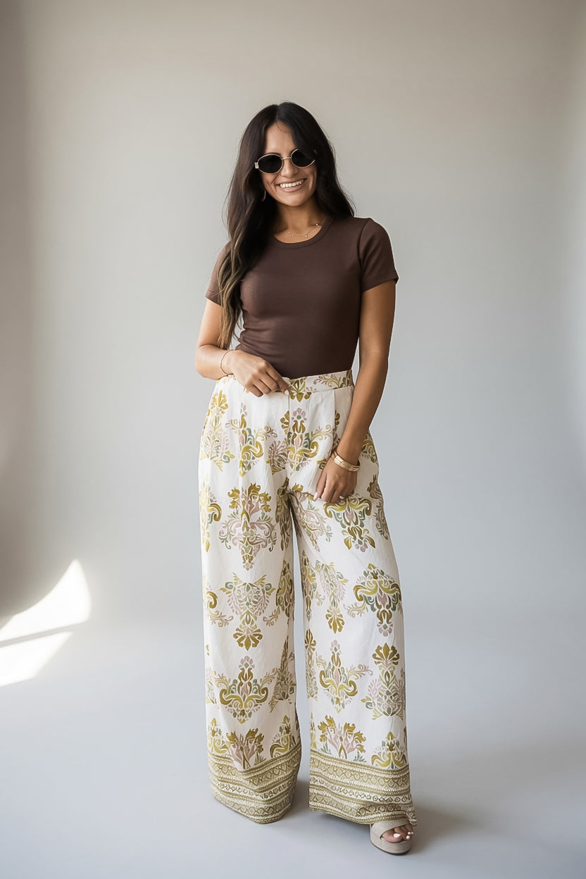 A woman modeling a brown short-sleeve crewneck top and wide-leg pants with an off-white base and olive green ornate pattern, standing against a neutral light gray studio background.