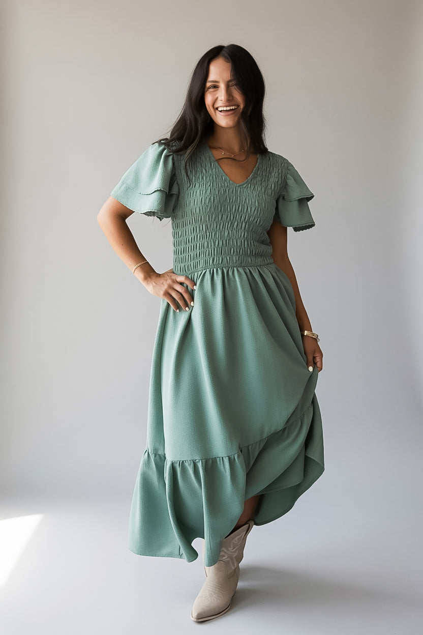 A woman modeling a sage green midi dress with flutter sleeves, a smocked bodice, and a tiered skirt, paired with beige ankle boots against a neutral studio background.