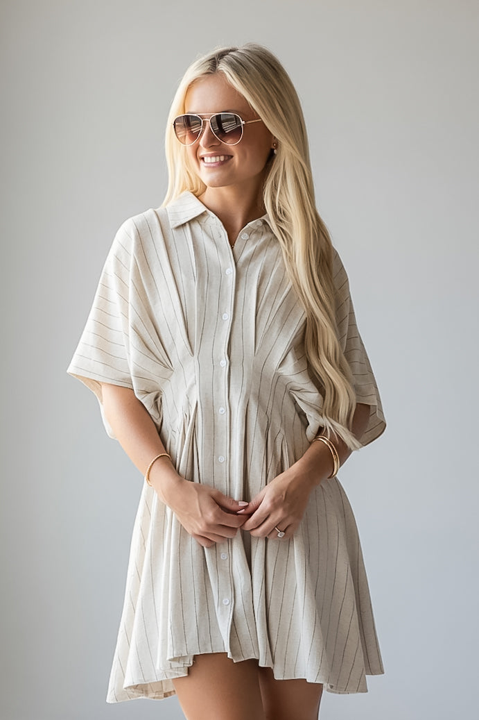 A blonde woman wearing a short-sleeve, button-front mini dress with vertical pinstripes in a light cream/beige color, styled with aviator sunglasses against a plain light gray studio background.
