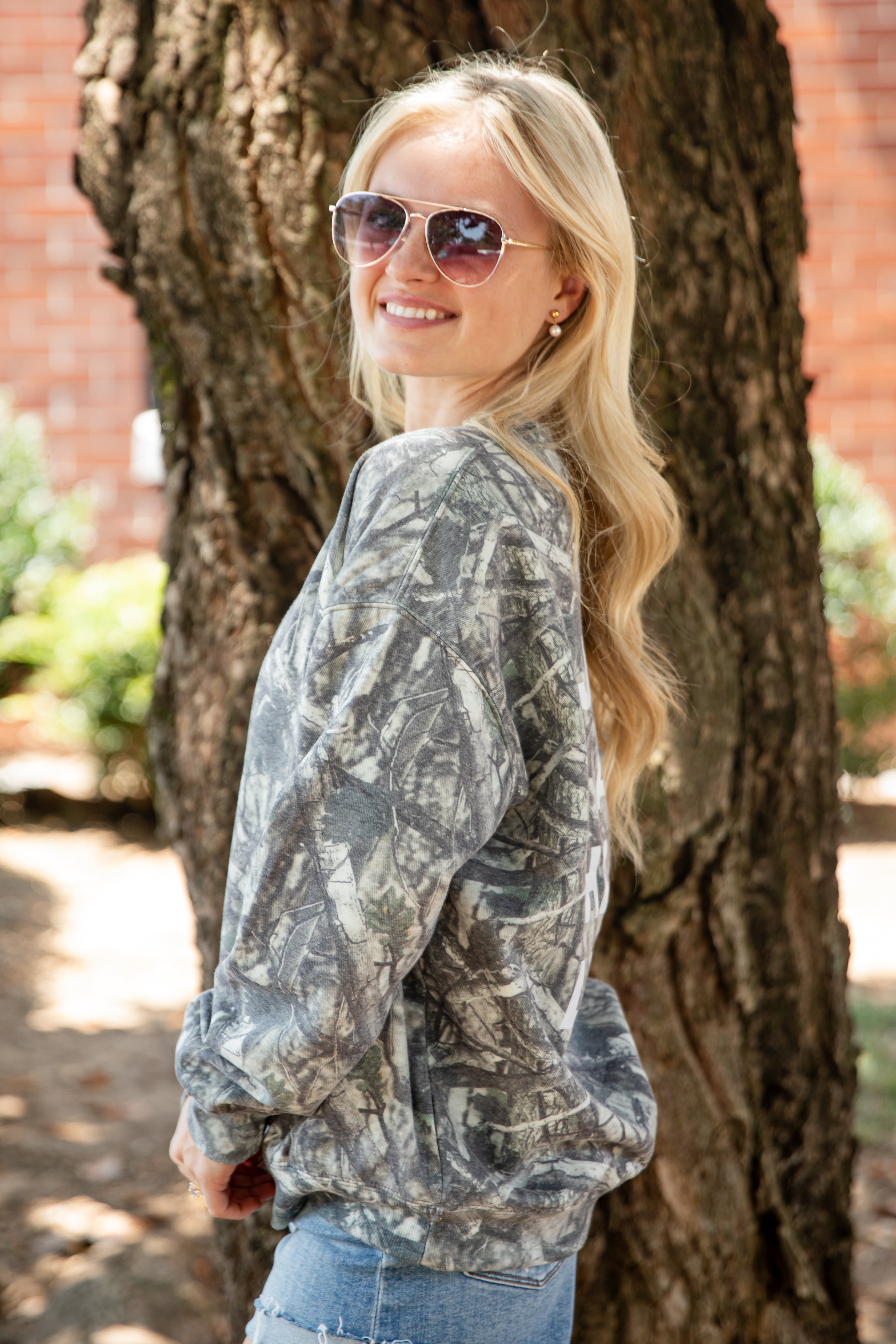 A woman with blonde hair and aviator sunglasses, wearing a gray and white camouflage pattern sweatshirt, posing outdoors next to a large tree trunk.