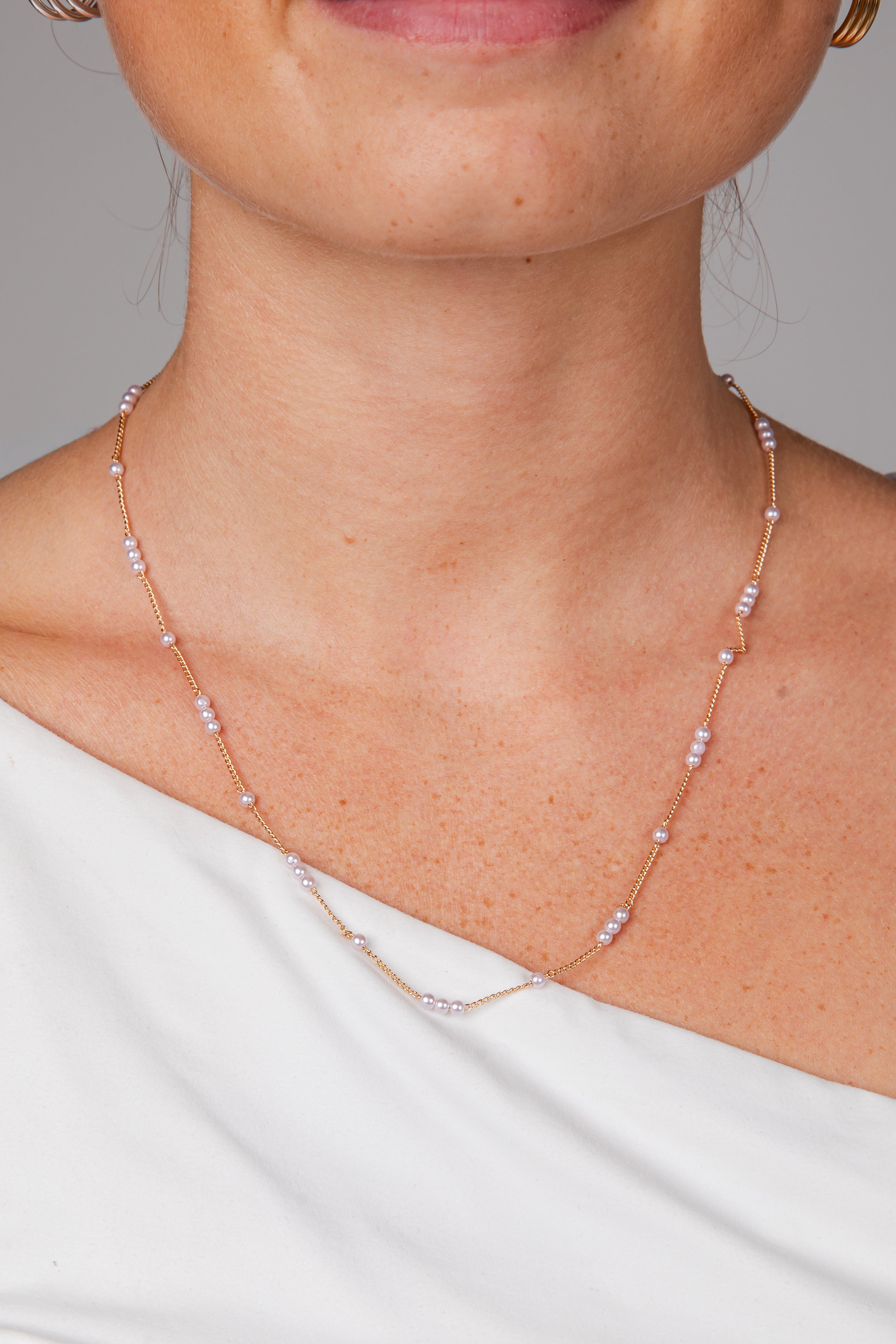 A close-up of a model wearing a delicate rose gold chain necklace with small white pearl-like beads spaced along the chain, paired with a white off-the-shoulder top.