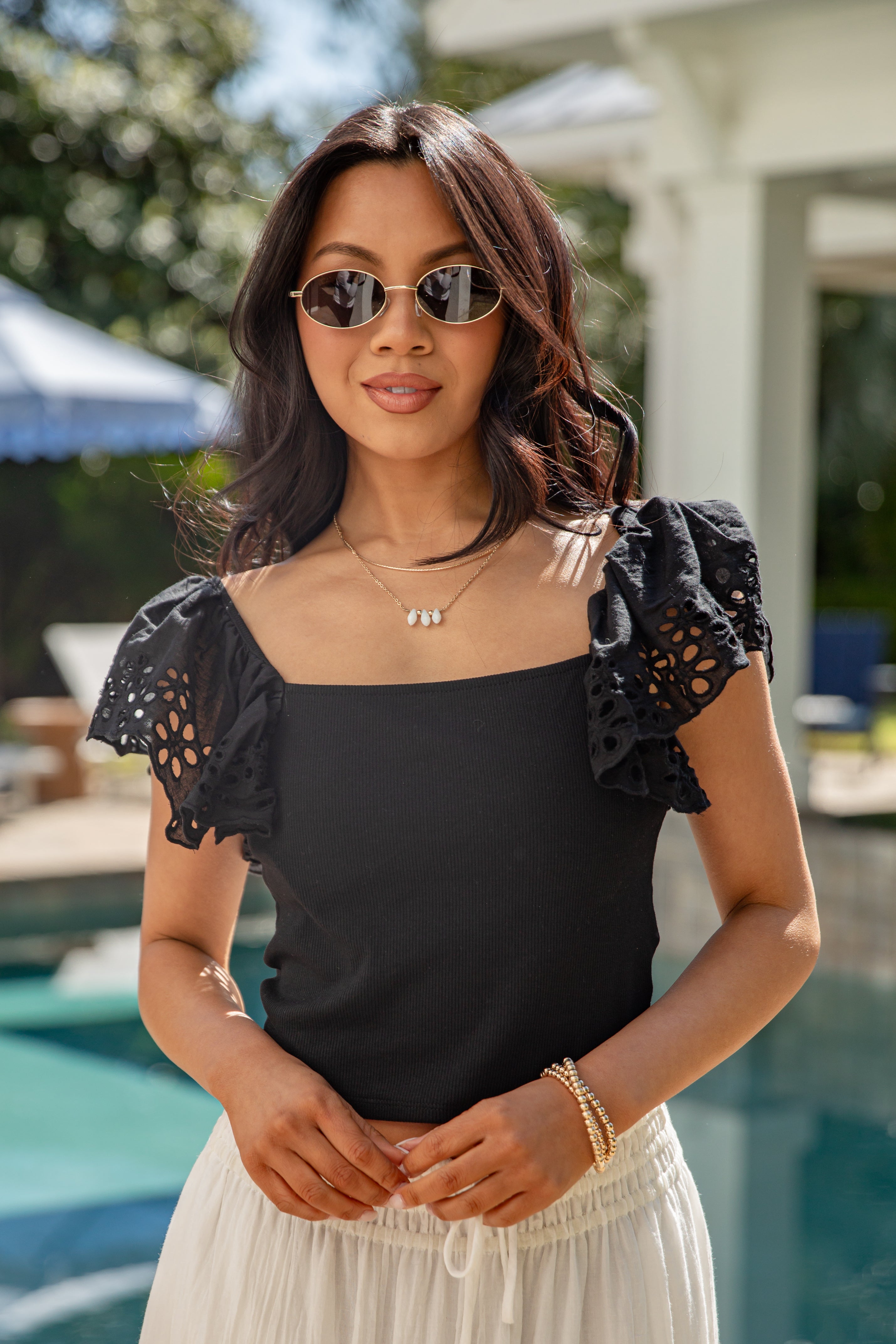 Woman in black eyelet-sleeve top and cream skirt by pool.