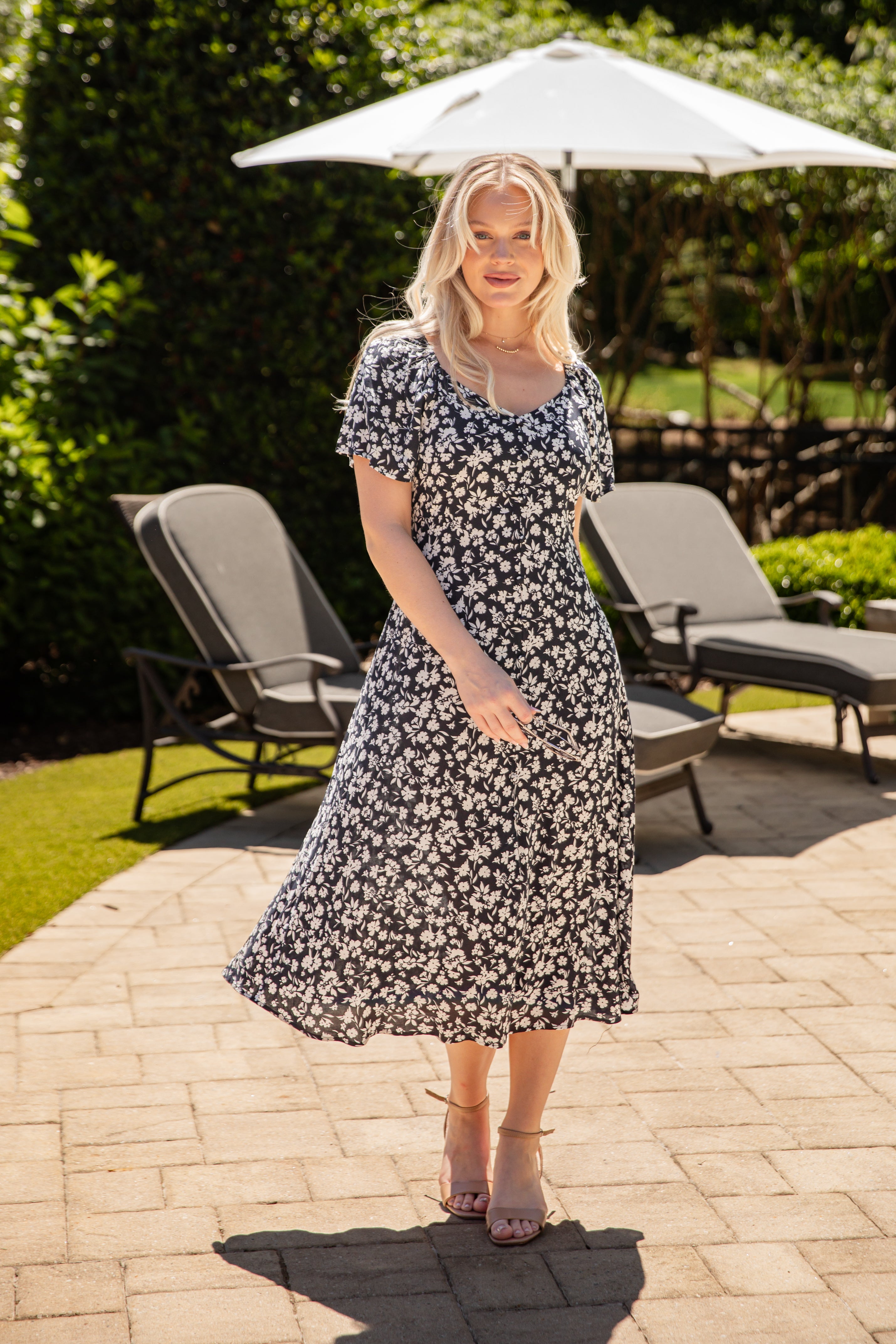 Woman in a floral dress standing on a patio with outdoor furniture and greenery in the background