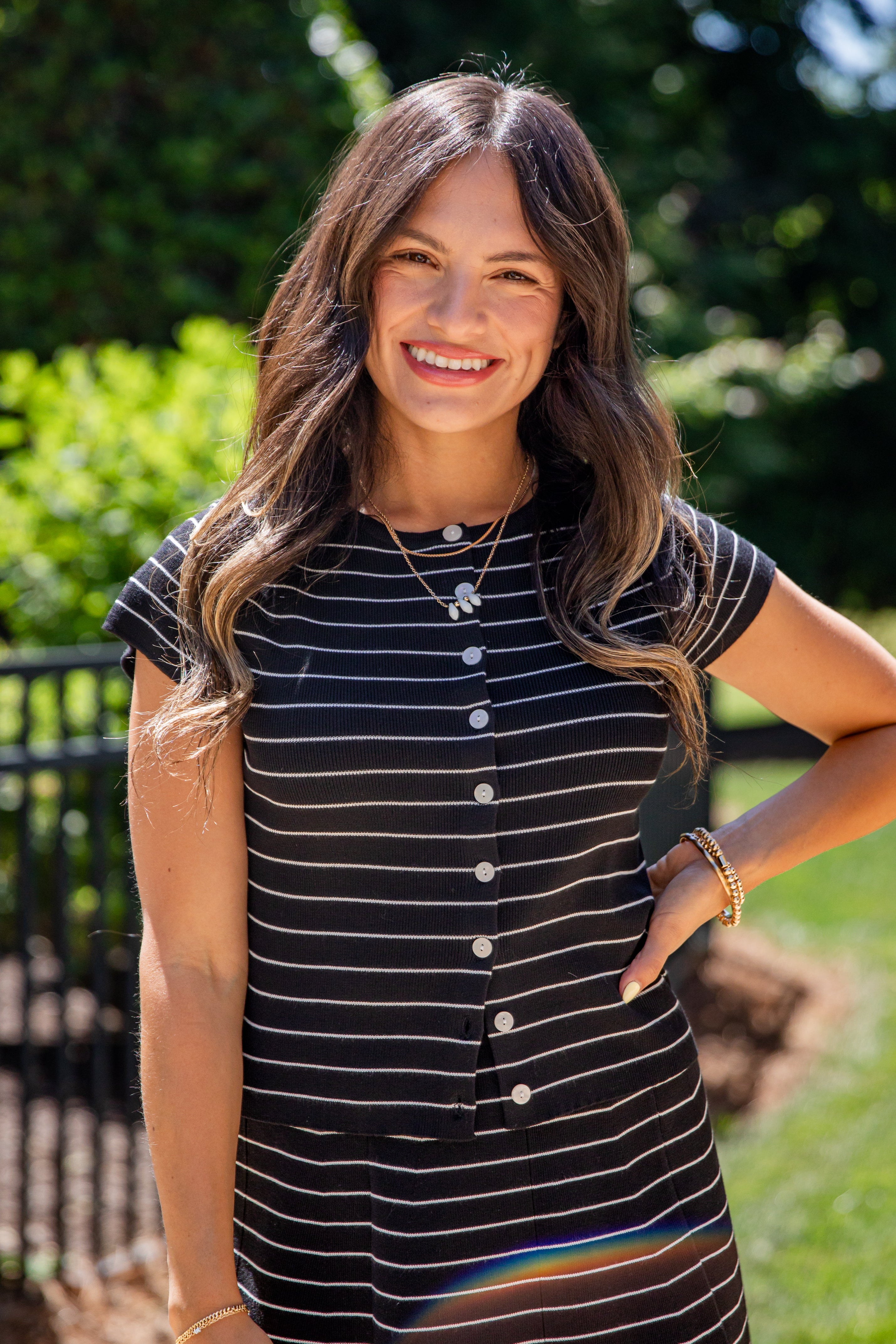 A woman smiling outdoors wearing a short-sleeve black and white striped button-front top , with green foliage and a fence in the background.