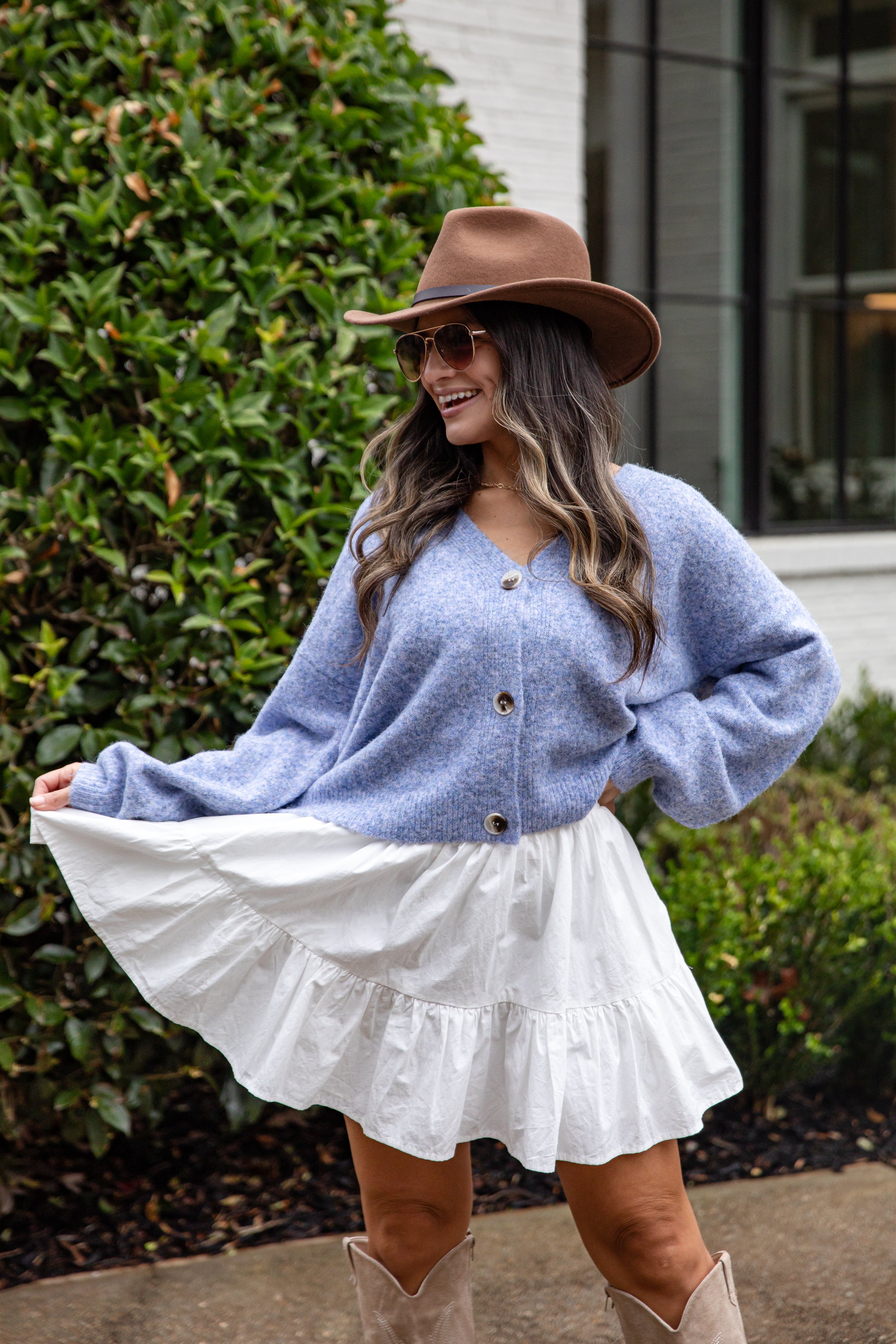 Woman wearing a blue cardigan, white skirt, and brown hat outdoors.