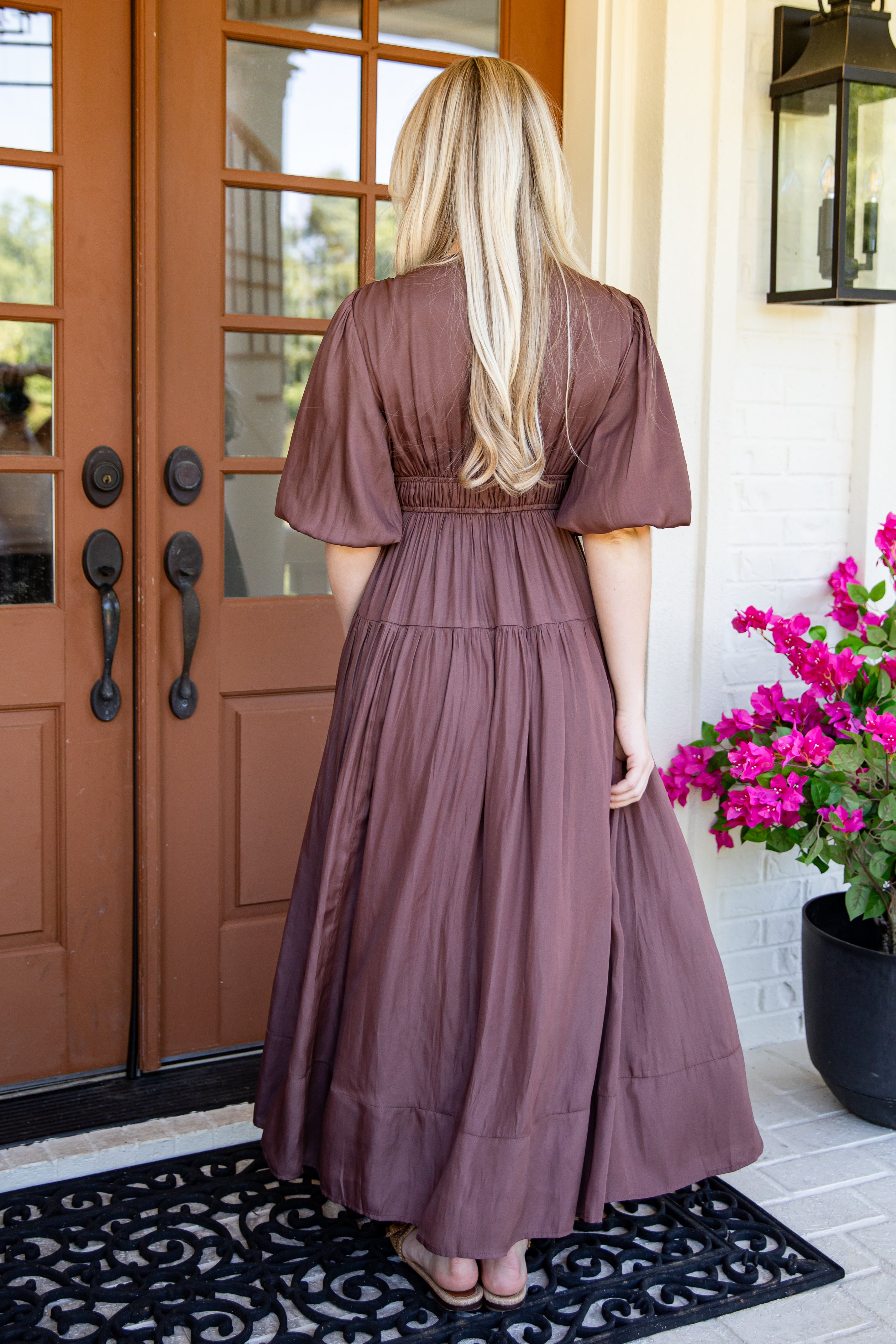 A woman stands with her back to the camera wearing a dusty mauve-brown tiered maxi dress with short puff sleeves, standing in front of a brown wooden front door with a black doormat and pink flowers on the porch.