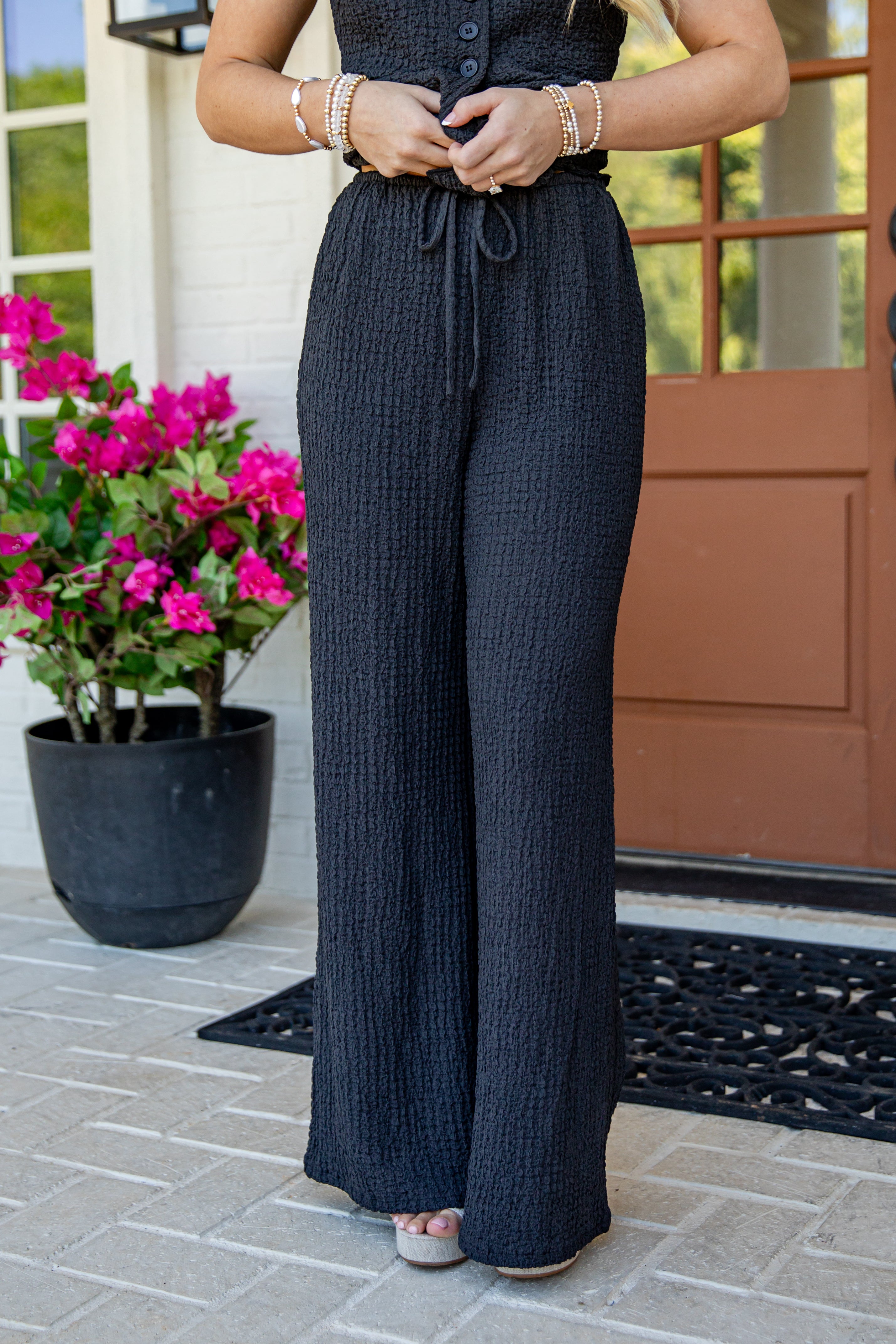A person wearing dark charcoal gray wide-leg textured pants with a drawstring waist, standing on a tiled porch with a potted pink flower and a brown door in the background.
