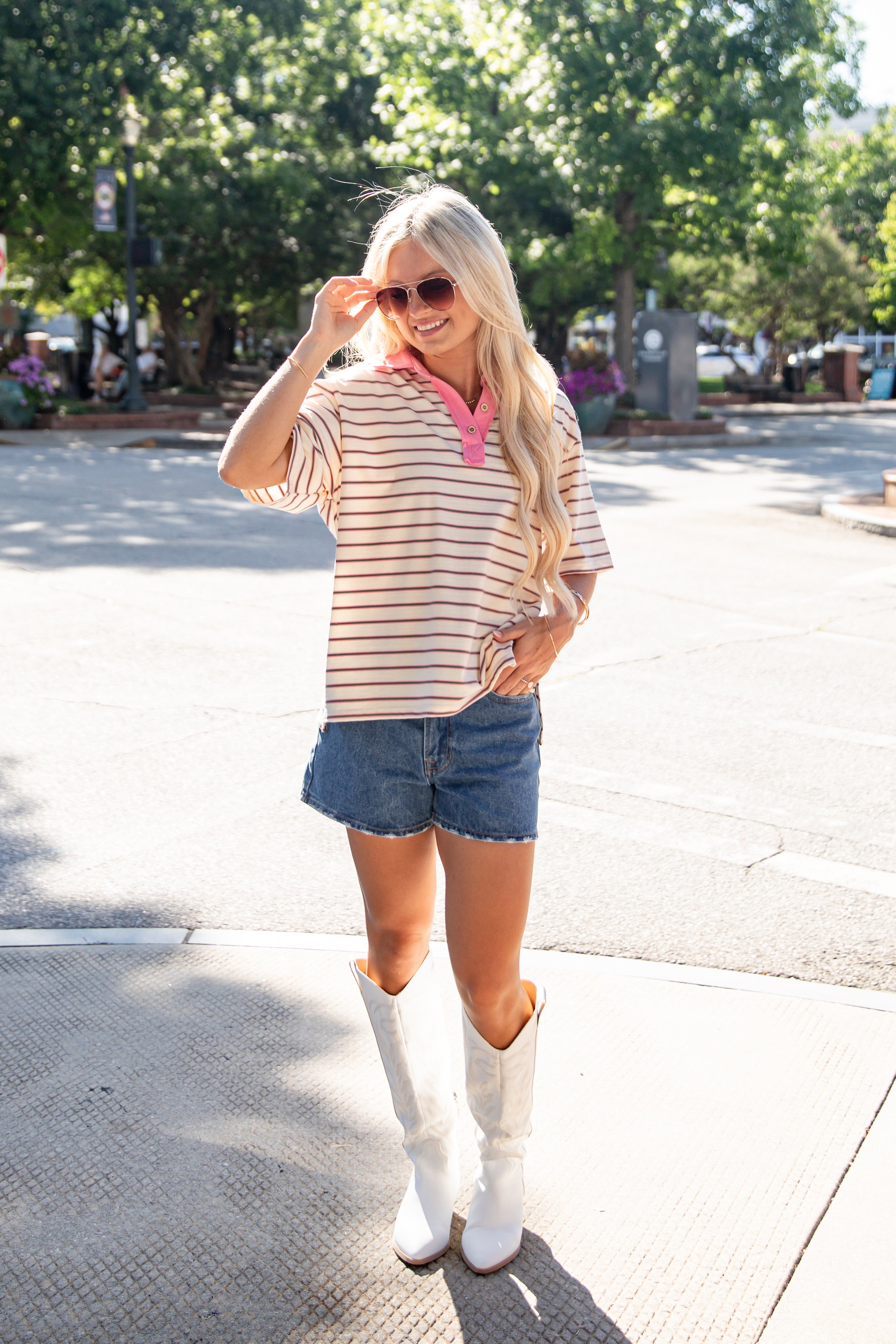 A woman stands on a sunny street wearing a cream and brown horizontally striped short-sleeve top with a pink collar, blue denim shorts, and white cowboy boots.
