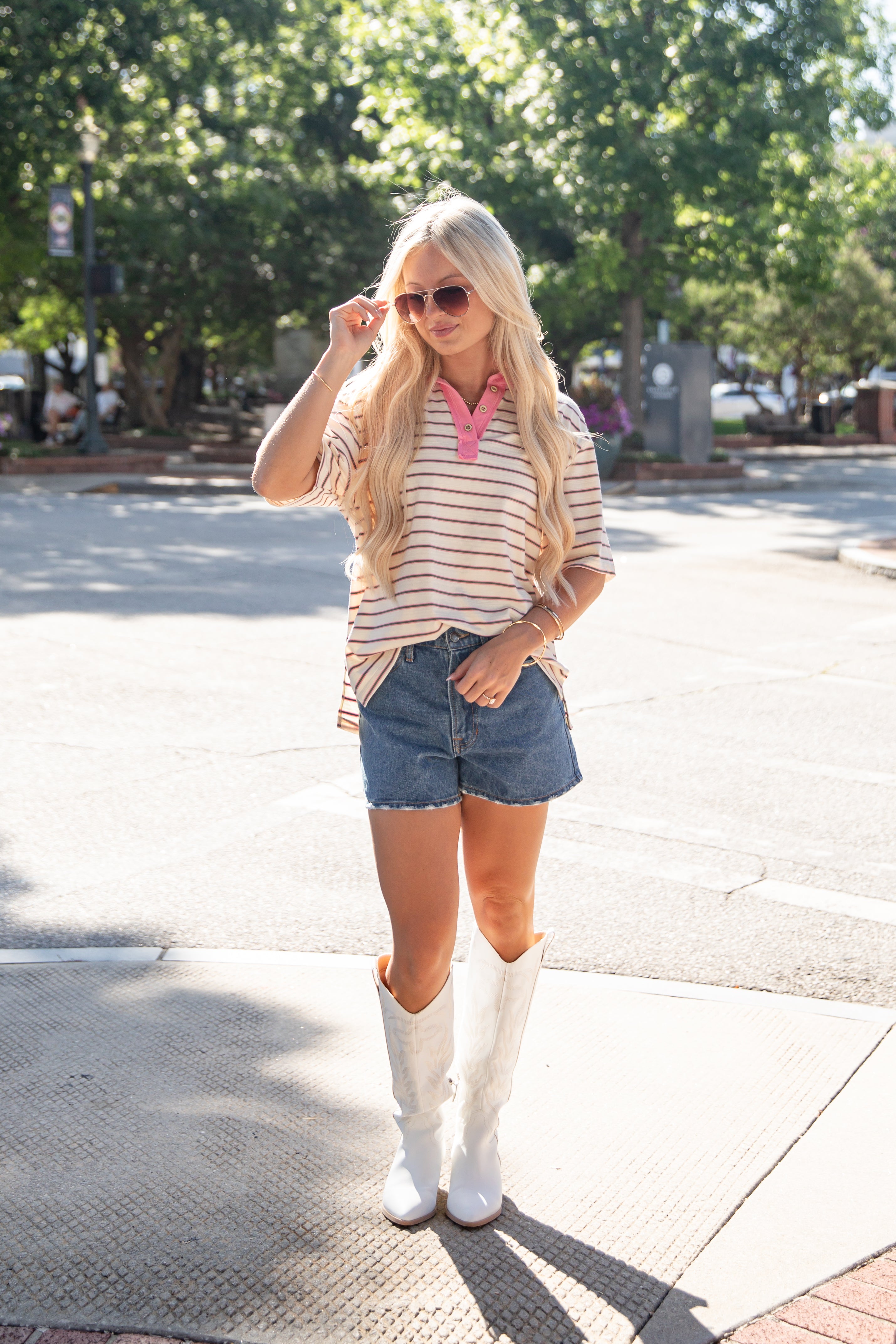 A woman stands outdoors on a sunny sidewalk wearing a cream and pink striped short-sleeve polo shirt, denim shorts, white cowboy boots, and sunglasses.