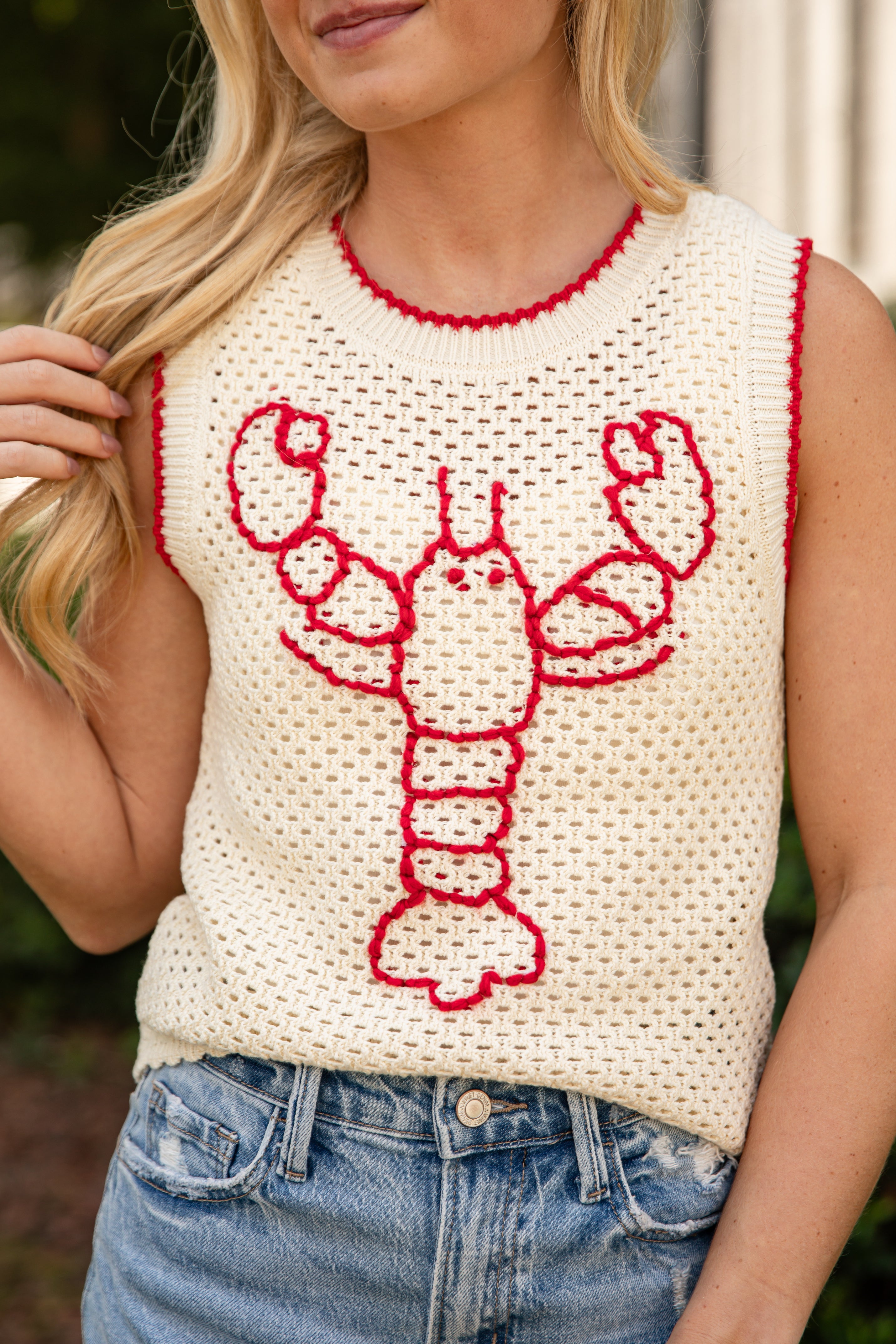 A woman wearing a sleeveless cream knit top with a red lobster graphic on the front, paired with blue jeans.