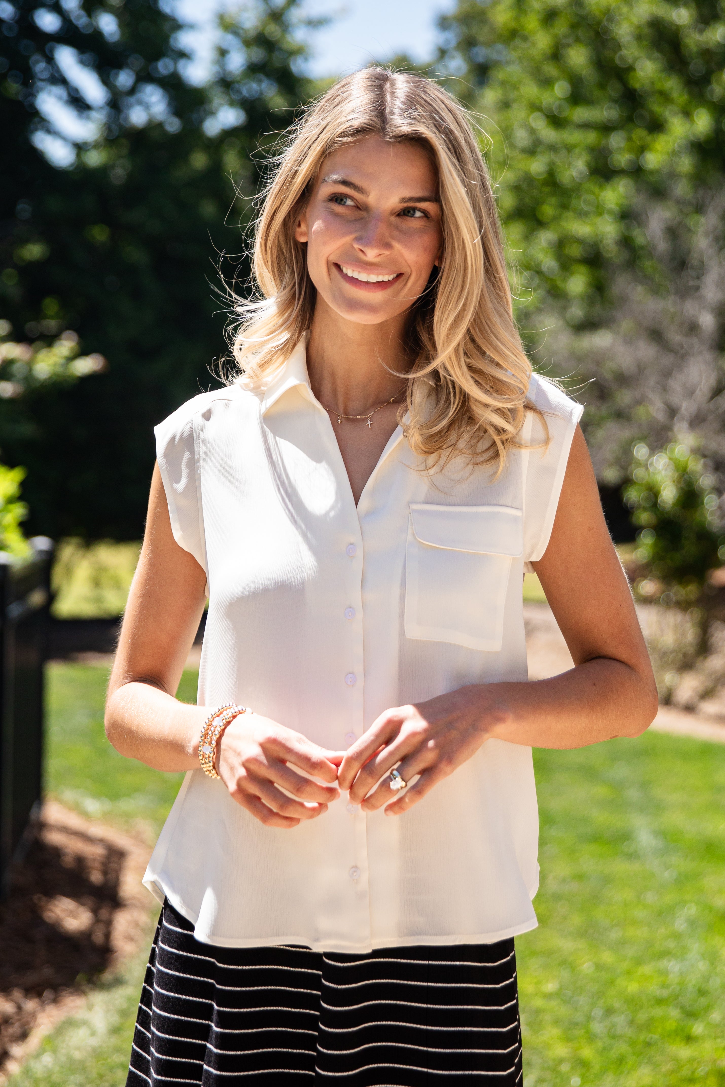 A woman wearing a sleeveless white button-up blouse and a black and white striped skirt, standing outdoors in a sunny garden setting.