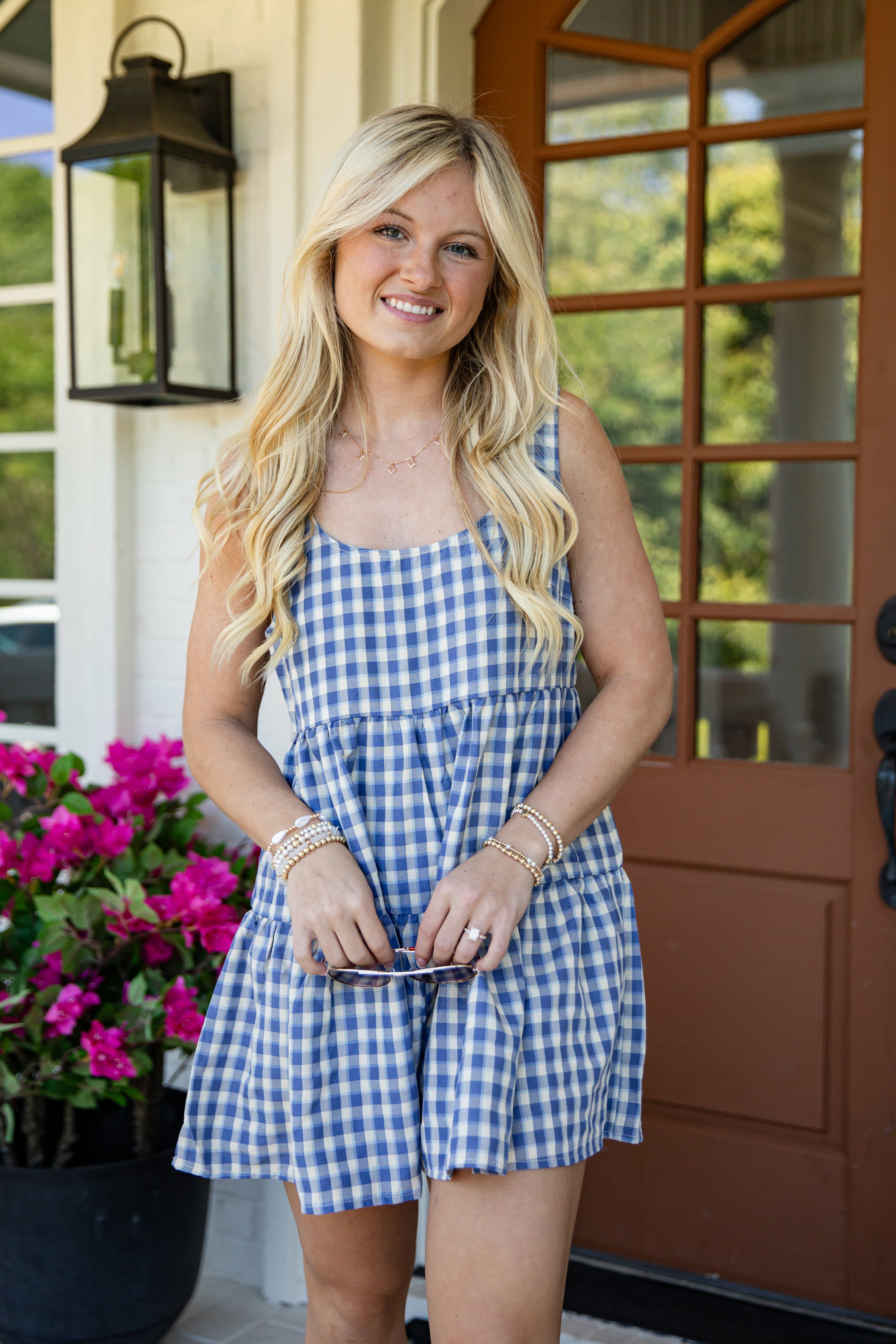 A smiling blonde woman wearing a blue and white gingham sleeveless romper stands on a front porch next to a brown door and pink flowers.