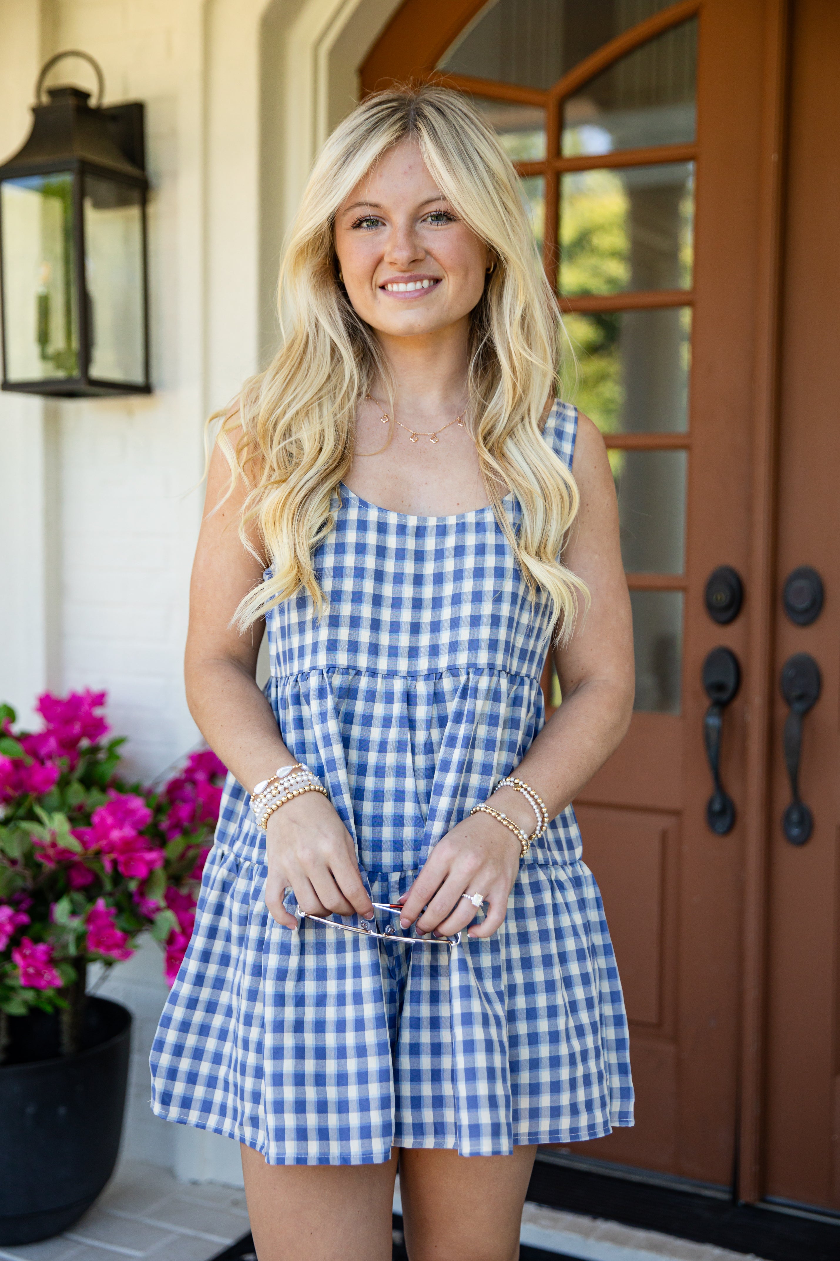A smiling blonde woman stands on a front porch wearing a blue and white gingham sleeveless romper. She is holding sunglasses, with a black lantern, a potted pink flower, and a brown wooden door in the background.