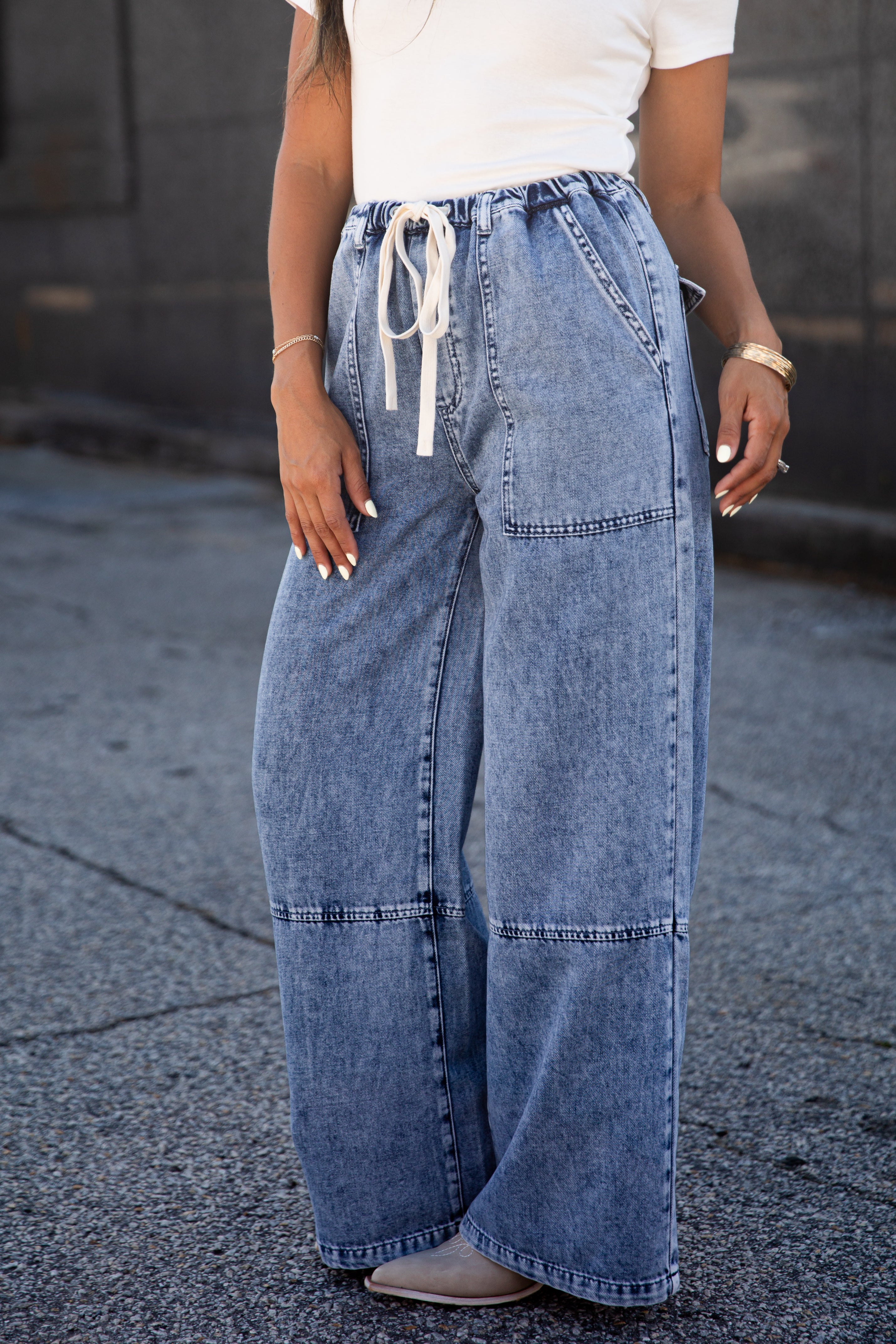 A person wearing wide-leg light blue denim jeans with a white drawstring waist, paired with a white top and gray shoes, standing outdoors on a paved surface.