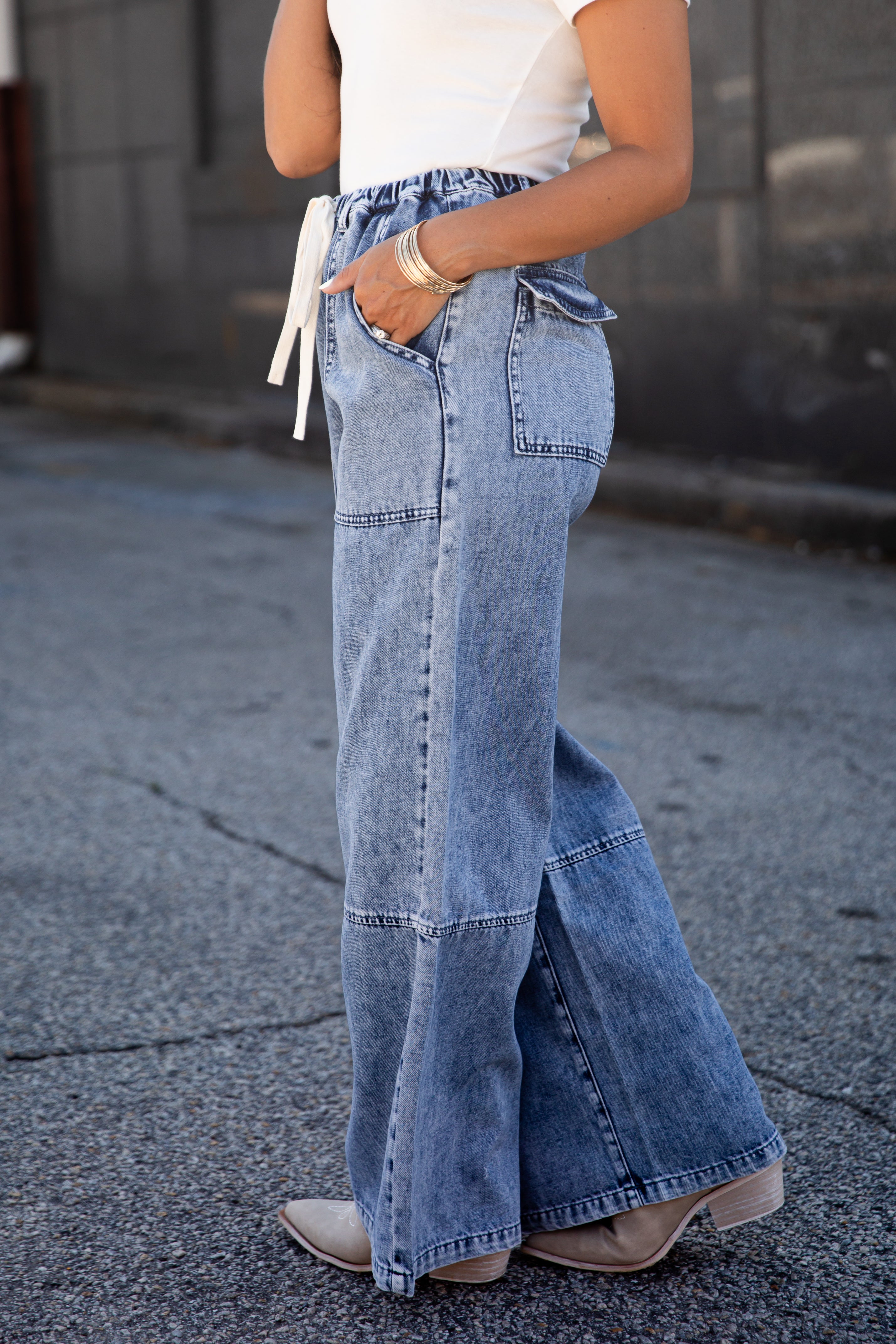 A person wearing wide-leg light blue denim jeans with a white drawstring waist, paired with a white top and beige ankle boots, standing on a paved street.