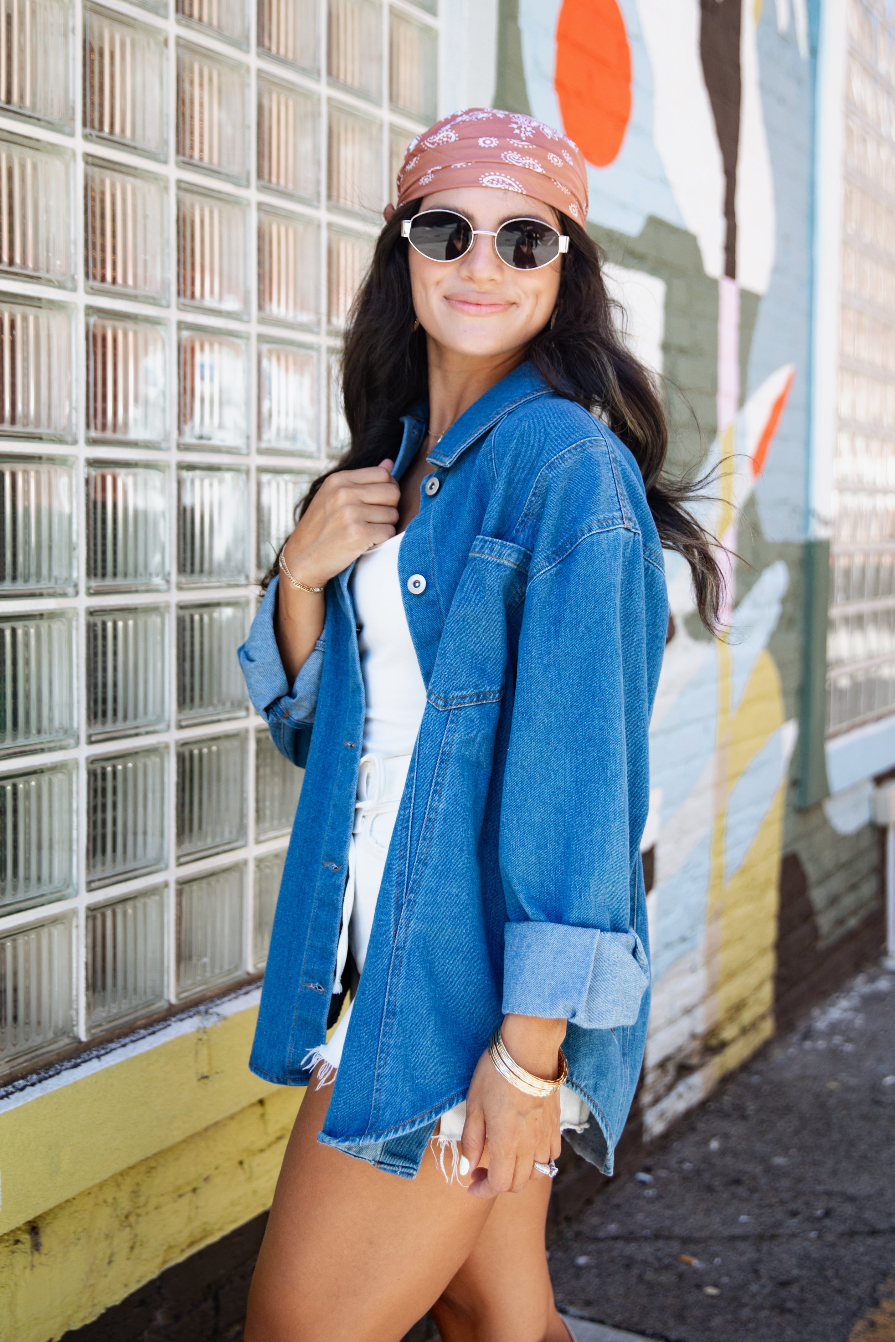A woman wearing a blue denim jacket over a white top, styled with a pink patterned bandana, sunglasses, and bracelets, standing in front of a glass block wall and a colorful mural.