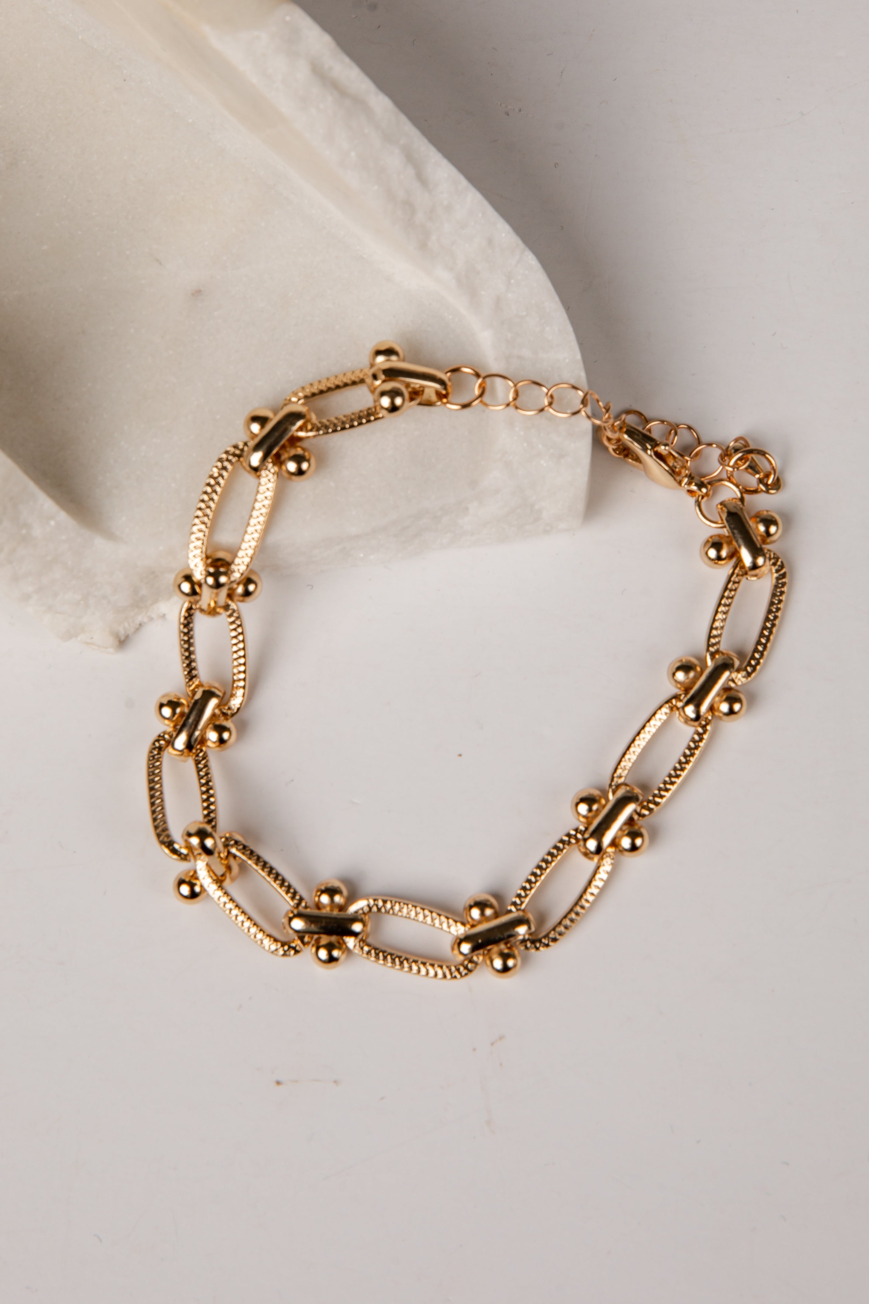A gold-toned metal chain bracelet with elongated oval links, small bead accents, and a short extender chain, displayed on a white surface next to a white textured block.