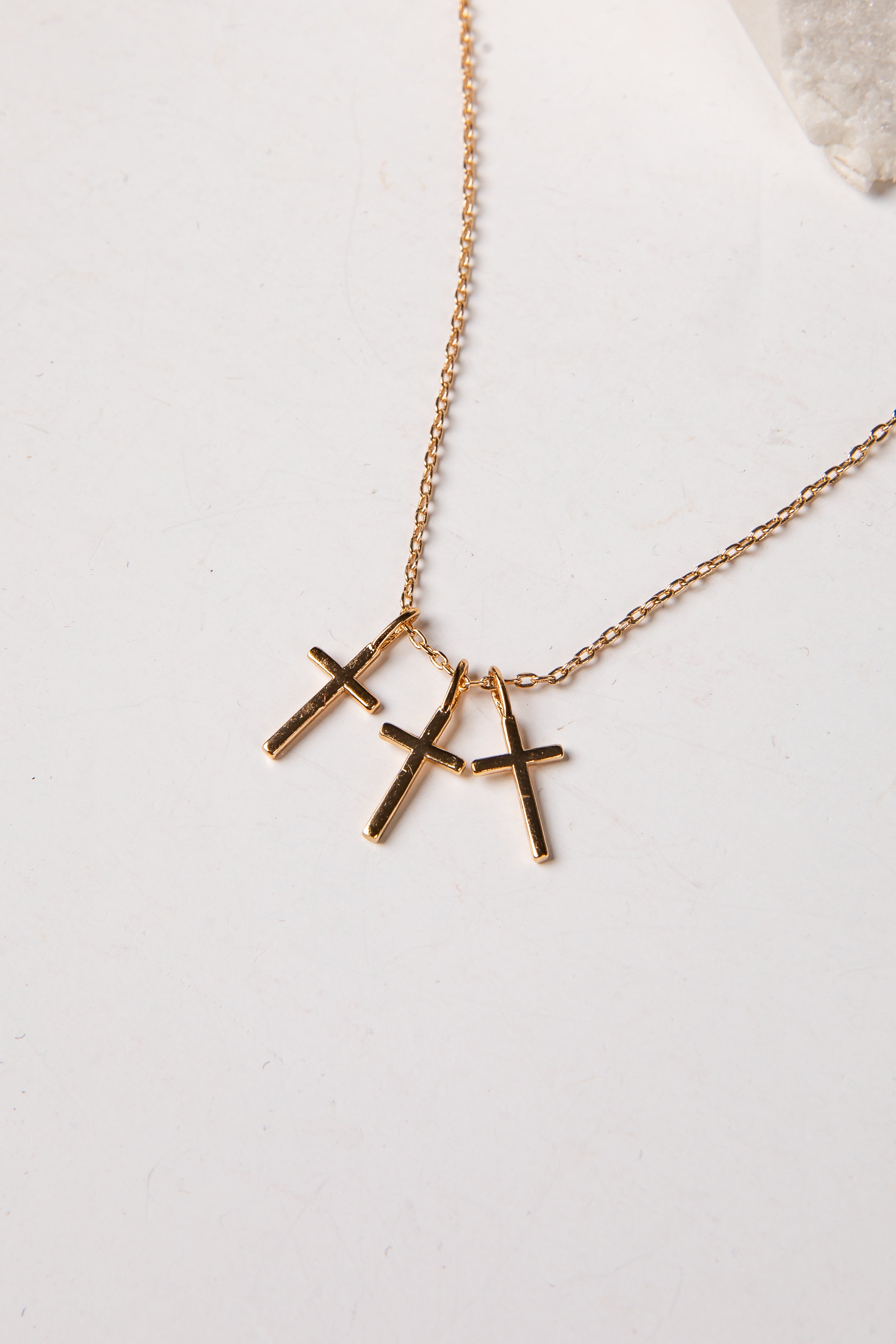 A gold chain necklace with three small cross pendants in a gold-tone finish, displayed on a light off-white surface.