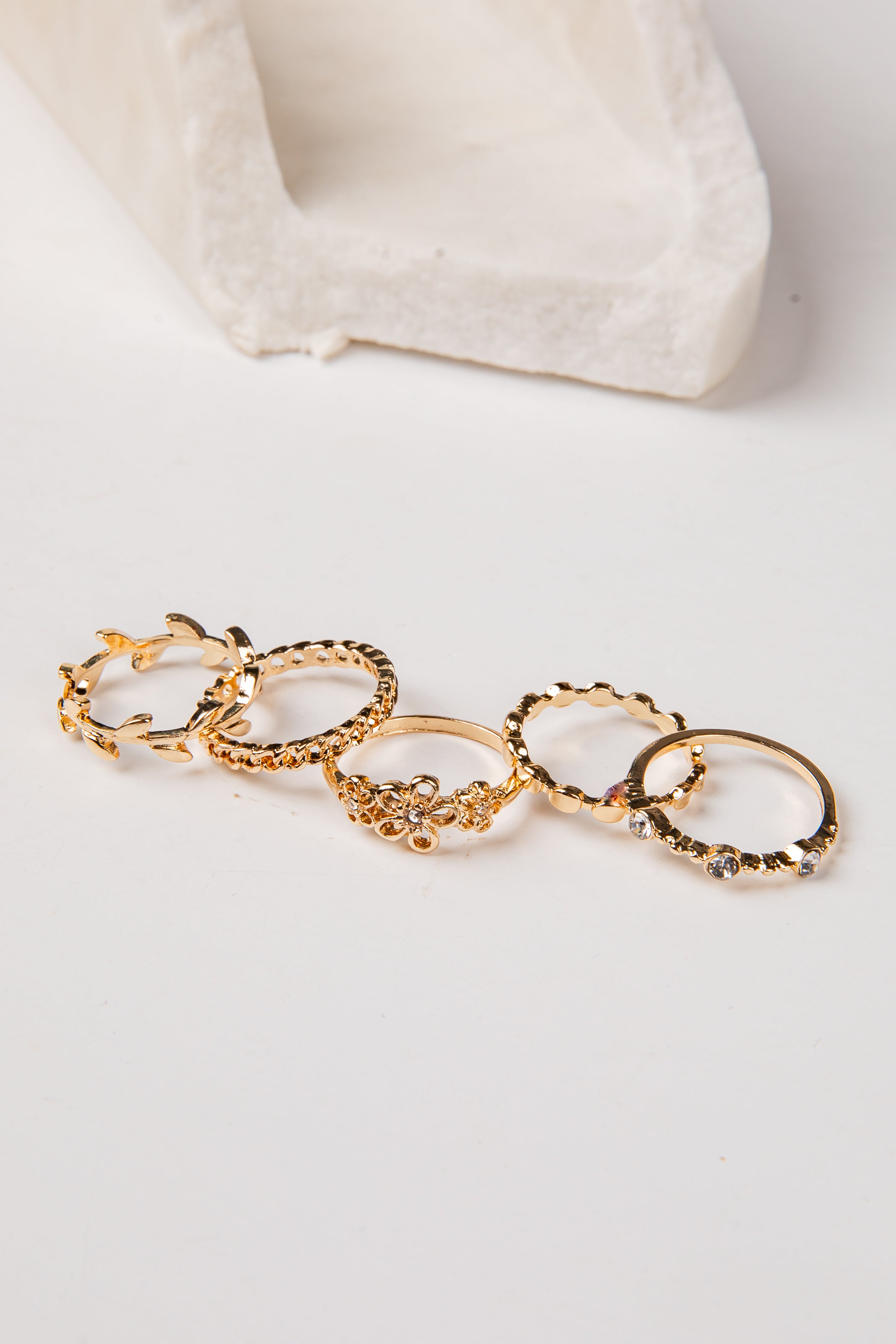 A set of five gold rings with textured, floral, and geometric patterns, arranged in a row on a white surface with a white textured block in the background.